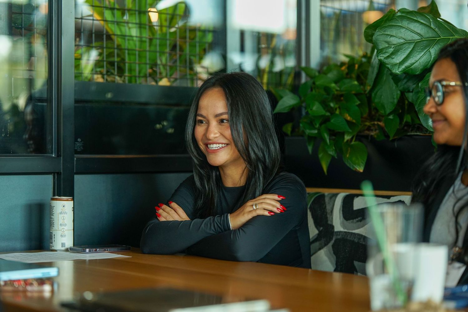 Smiling woman with crossed arms sitting at a cafe table surrounded by lush green plants.
