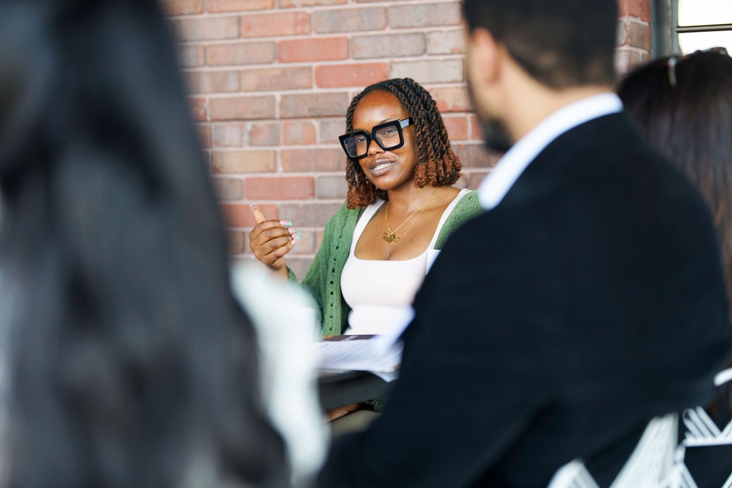 Young woman with glasses speaking confidently during a professional meeting near a brick wall.