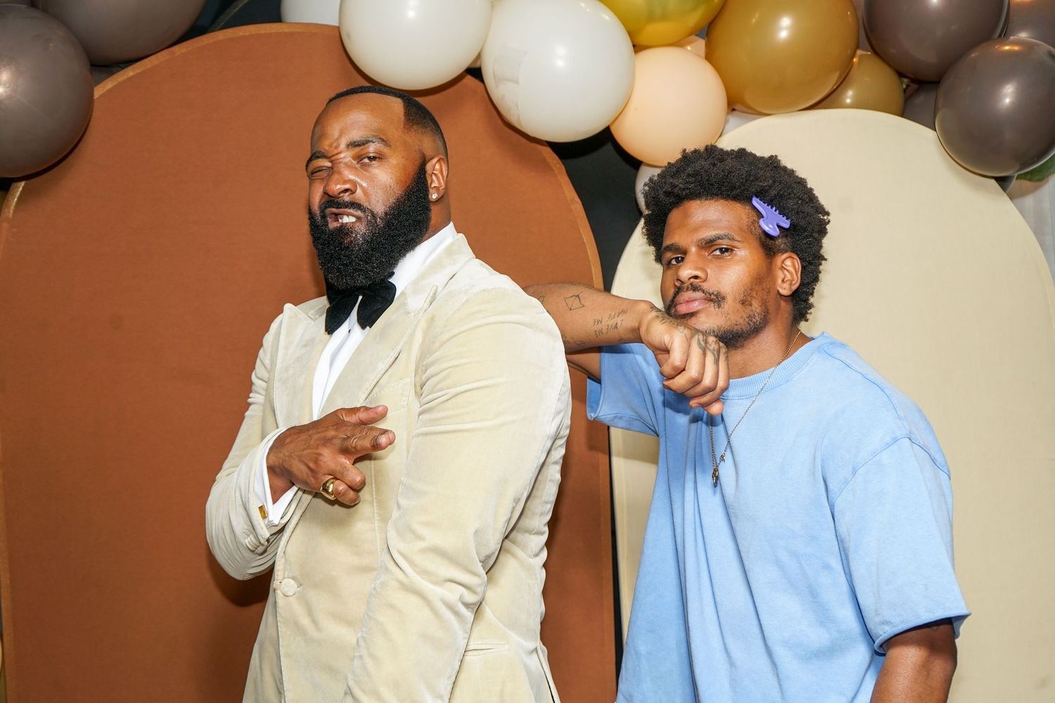 Two well-dressed men posing at a festive event with balloon decorations in neutral tones behind them.