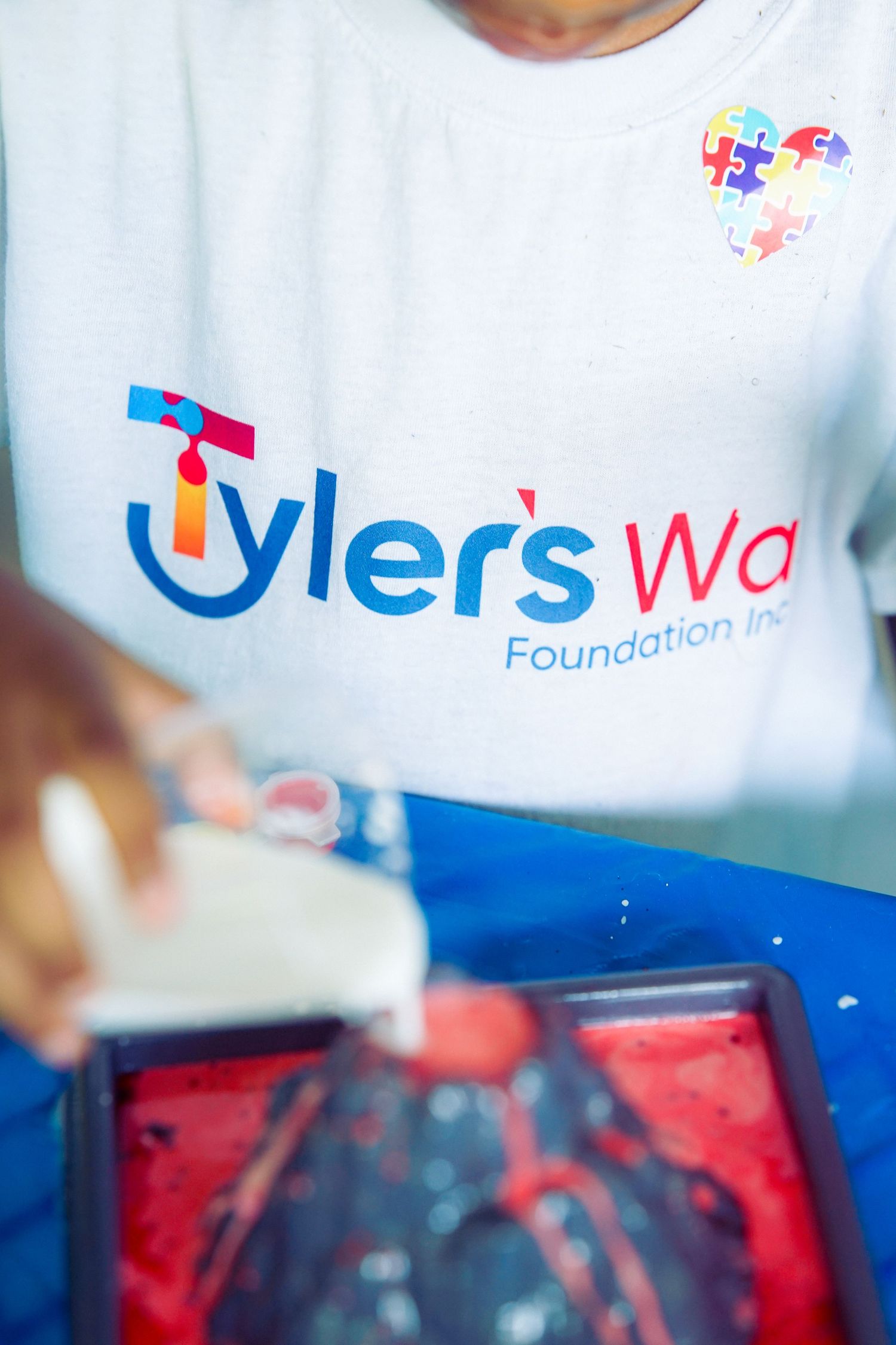 Person wearing Tyler's Way Foundation Inc. t-shirt using a squeegee with red paint on a screen printing tray.