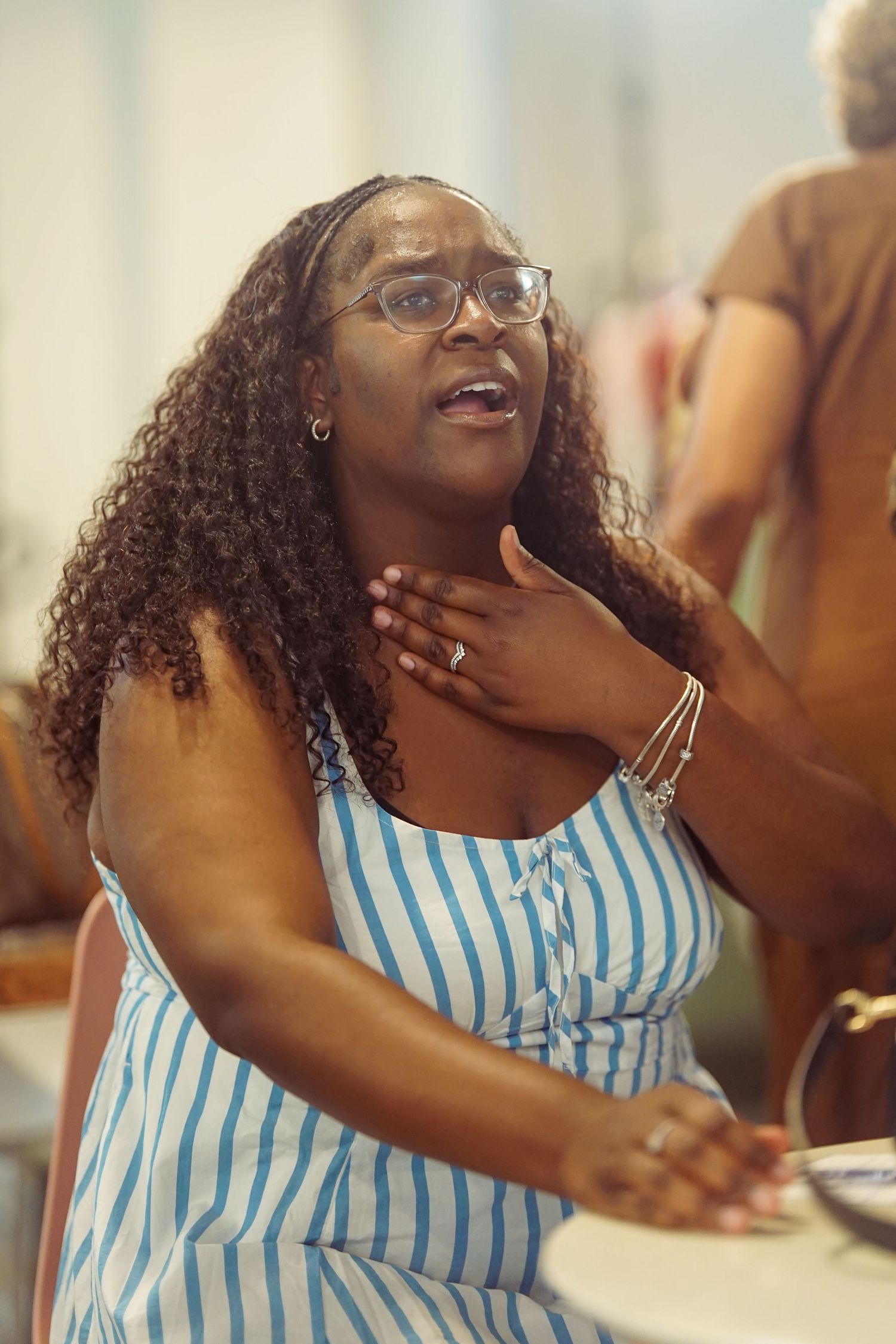 A Black woman in a striped dress and glasses places her hand on her chest while speaking emotionally.
