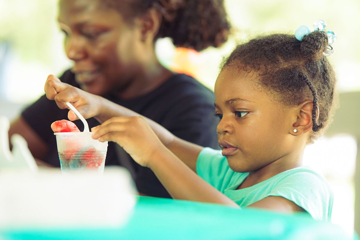Young Black girl stirs drink at outdoor community event while adult supervises nearby.