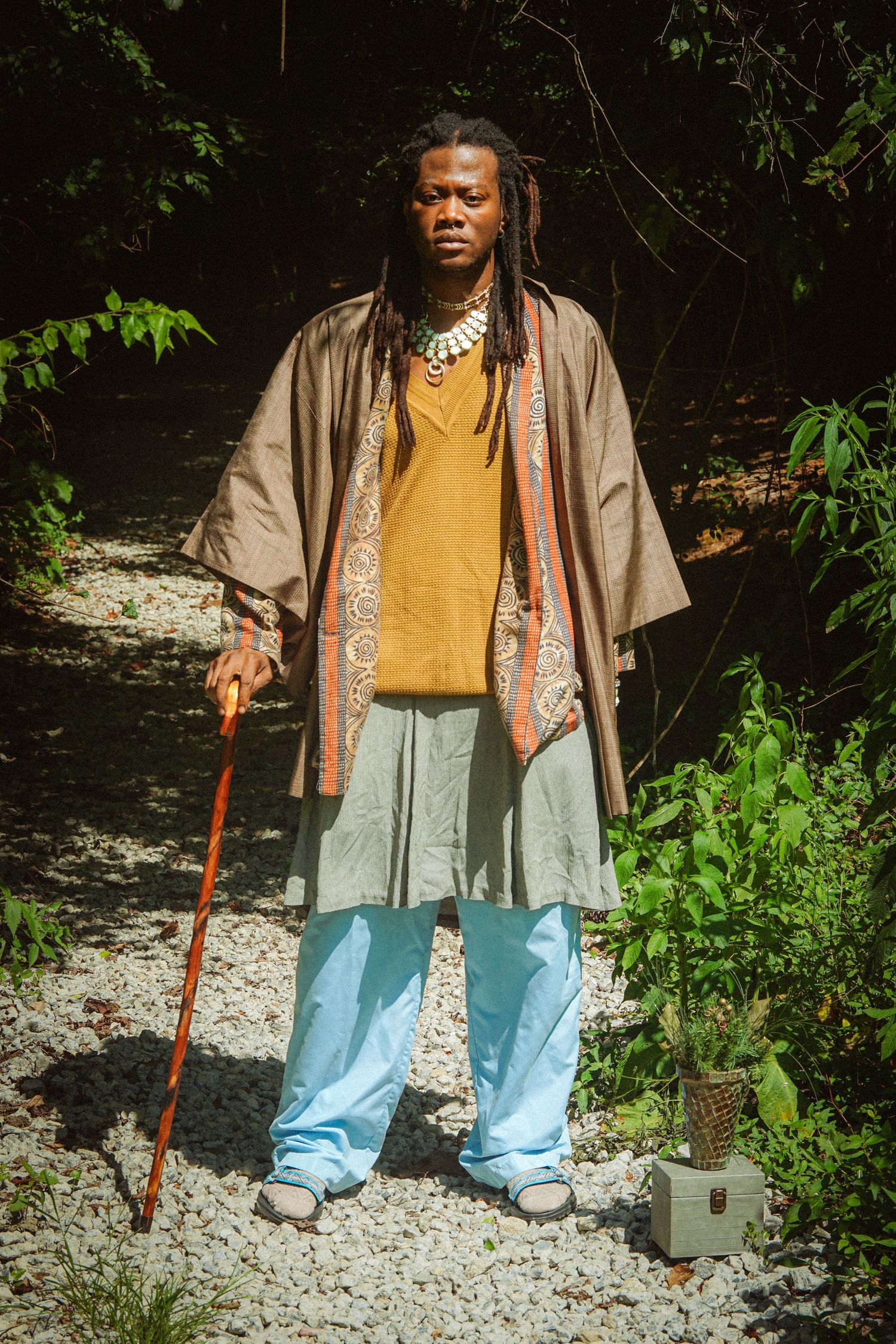 A man with dreadlocks in a brown kimono, yellow shirt, and light blue pants holds a wooden cane on a sunlit gravel path.