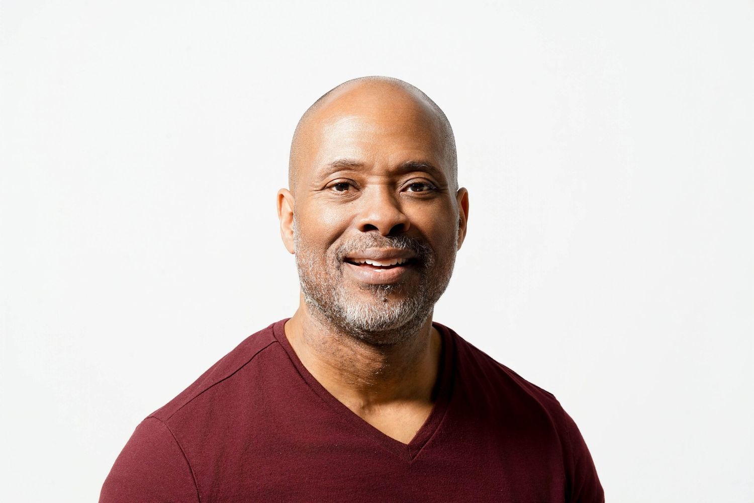 Smiling bald middle-aged Black man with gray beard wearing a dark red v-neck shirt against white background.