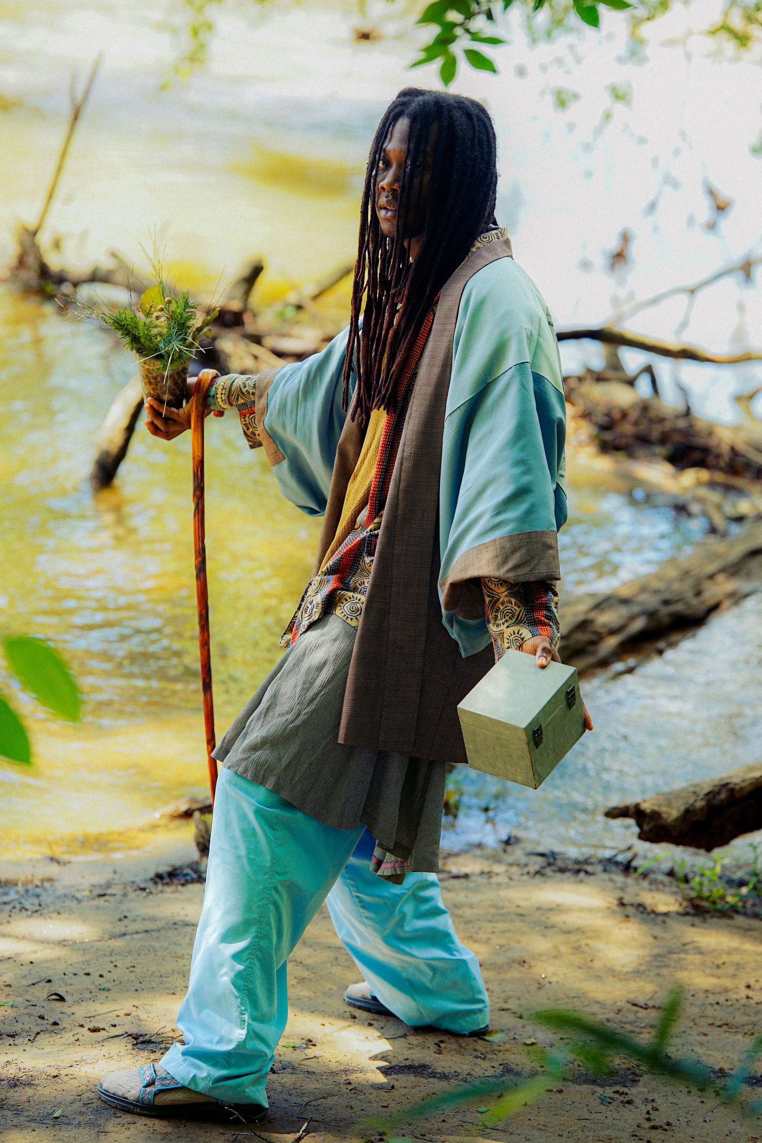 Person with dreadlocks in layered robes walks near water holding a wooden staff with plants and a small box.