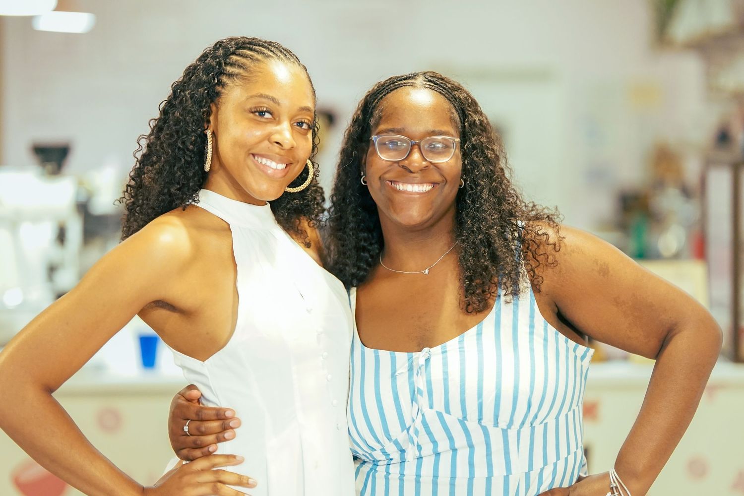 Two women smile confidently with hands on hips while posing together at an indoor event.