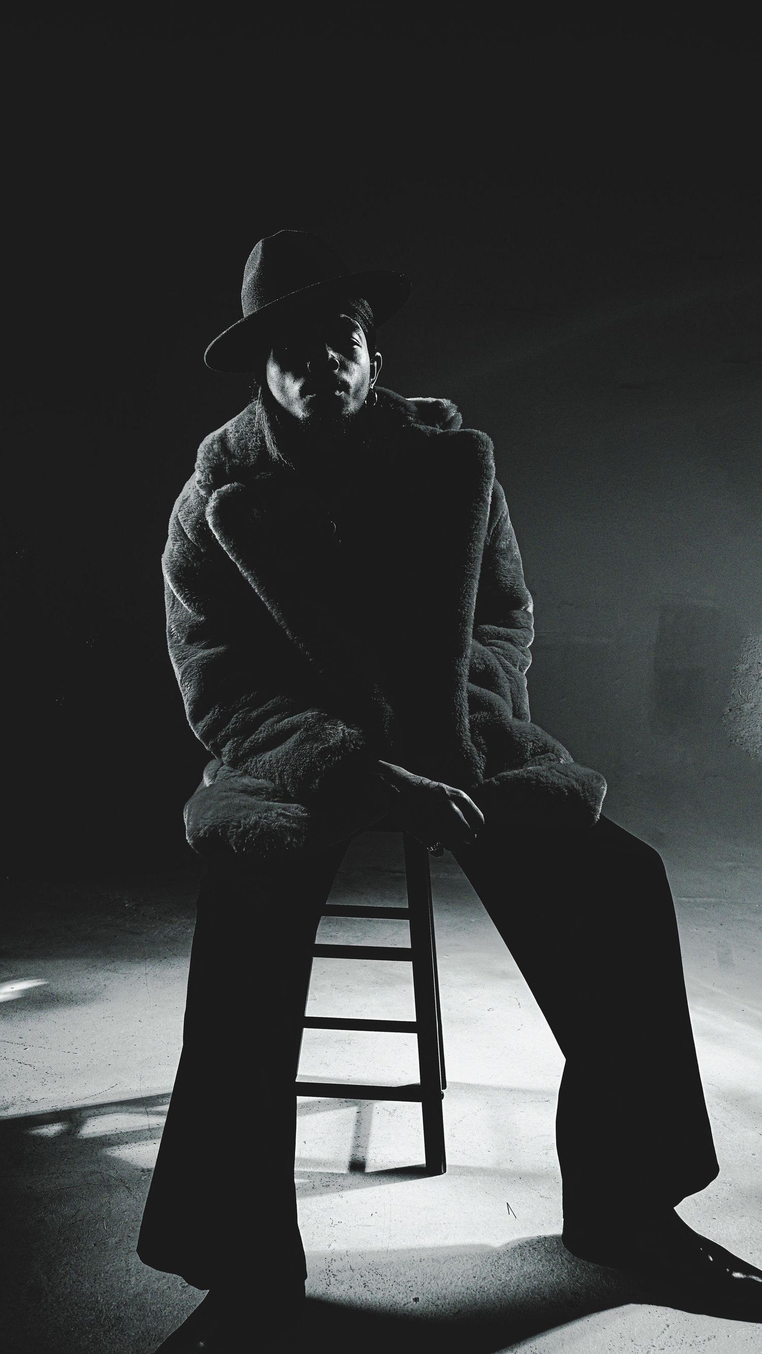 Moody black and white sequence showing a seated figure in shadow on a wooden stool.