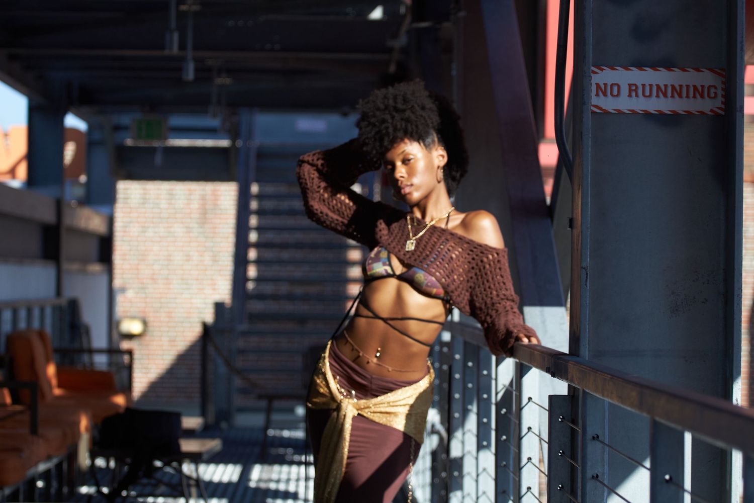 Model in brown knitwear and gold skirt poses dramatically in industrial setting with shadows and light.