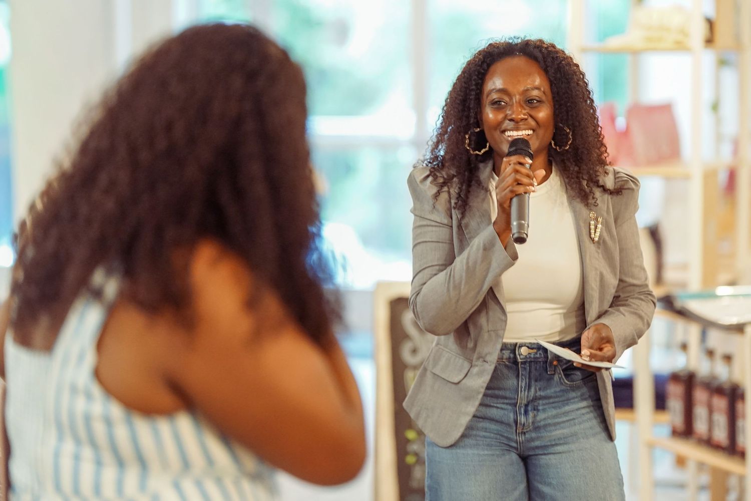 A confident Black woman speaks into a microphone at an indoor event, smiling warmly at the audience.