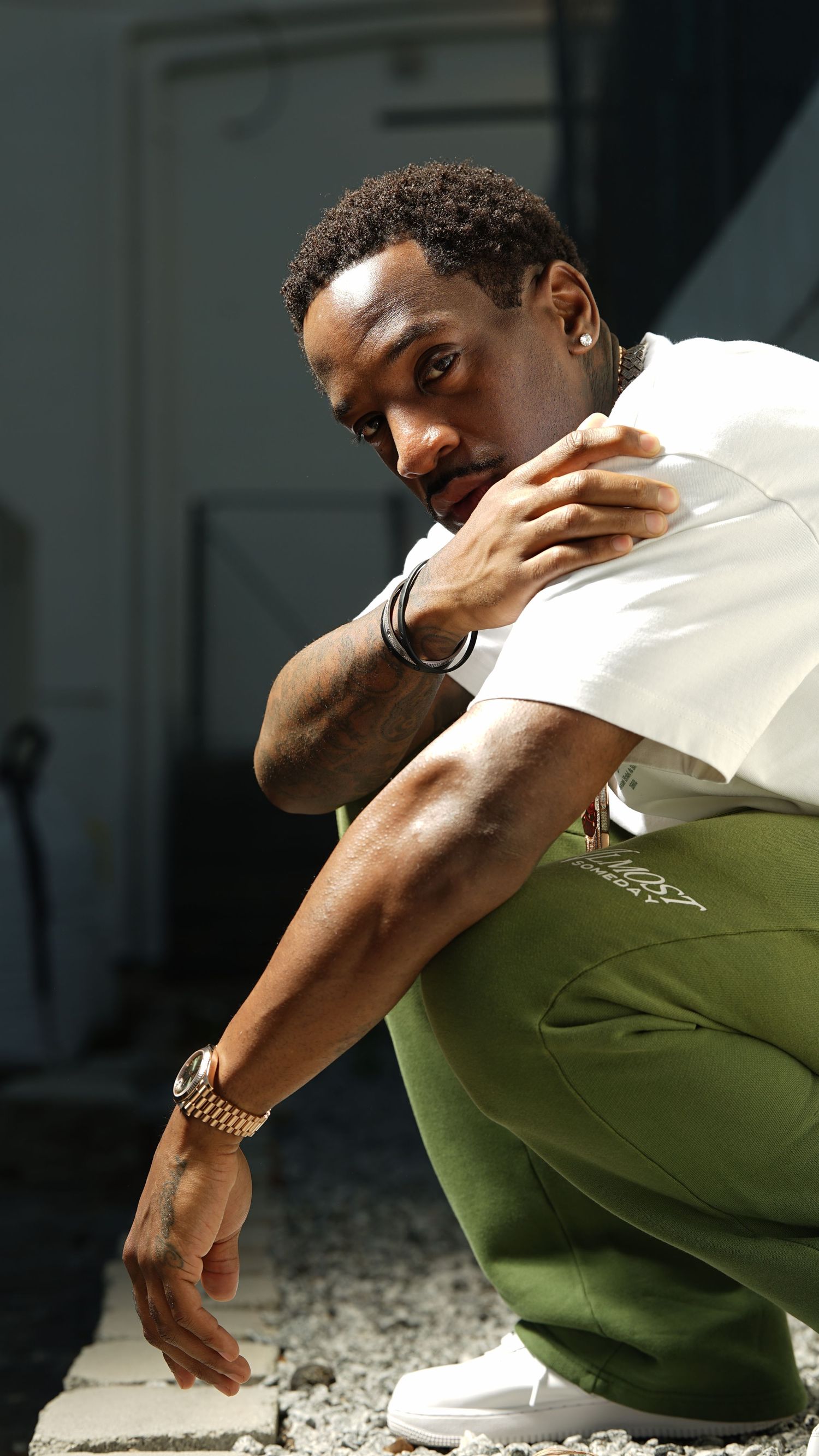 Person in white shirt and green pants sits in contemplative pose against dark background.