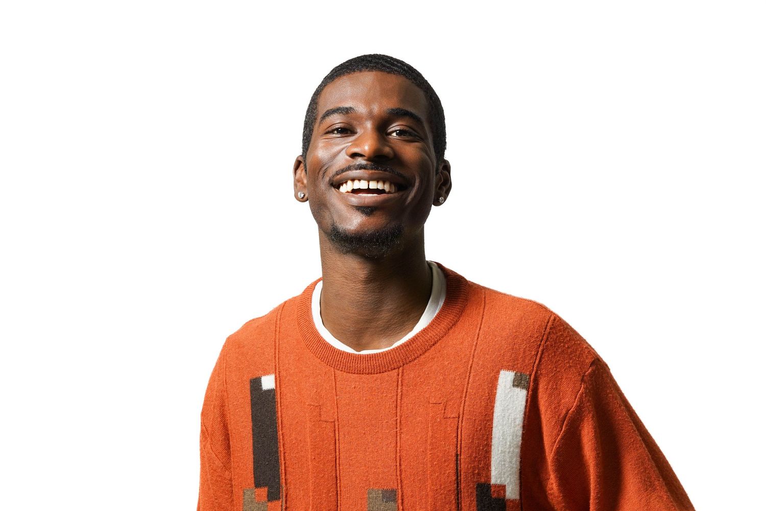 Smiling young Black man wearing an orange sweater with geometric patches, photographed against a white background.