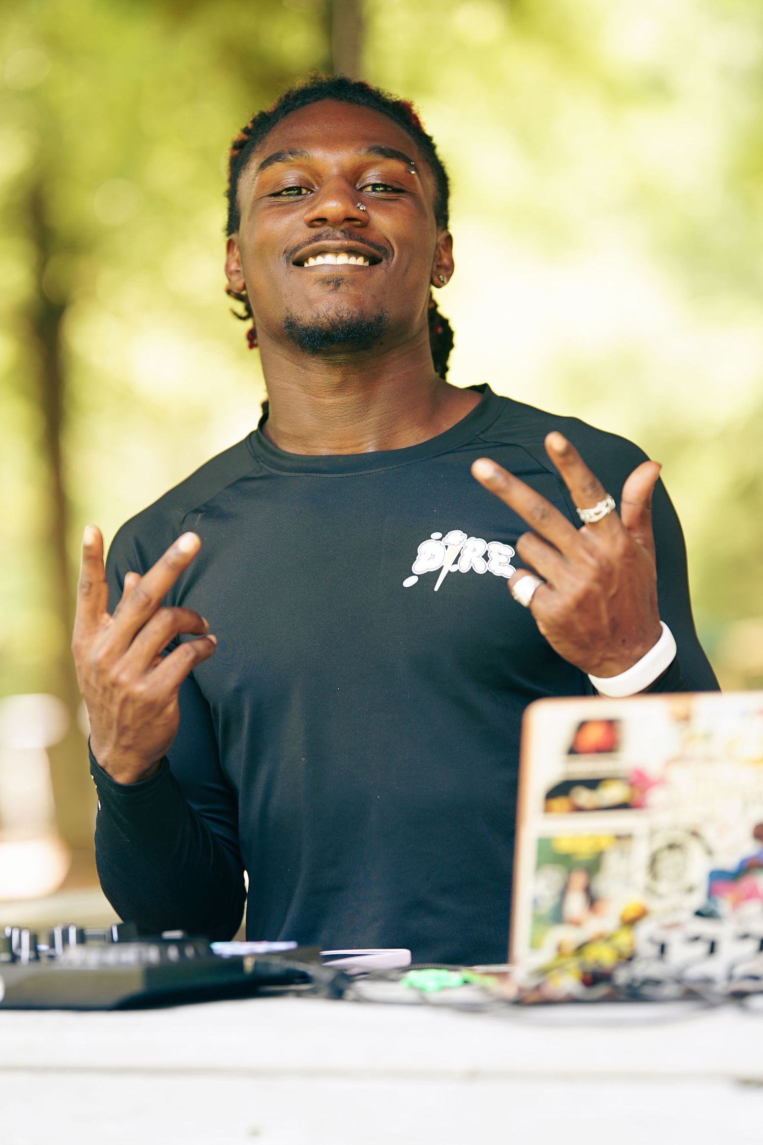 A smiling DJ performs outdoors at a turntable setup, flashing hand signs while wearing a black long-sleeve shirt.