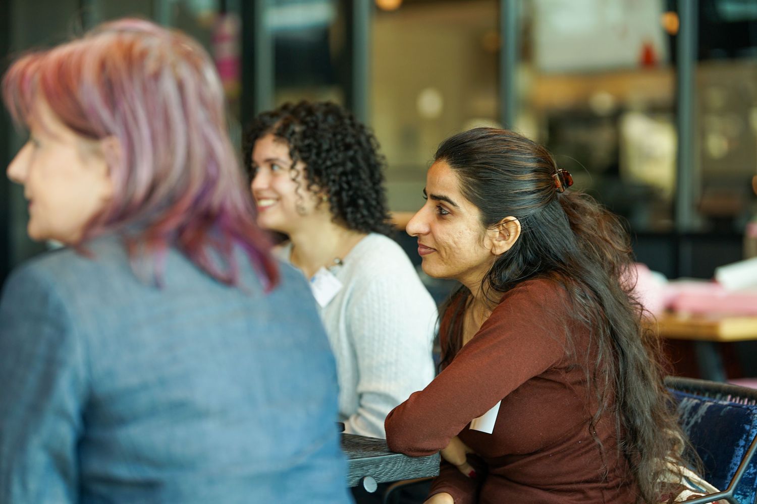 Three diverse women smile and engage in conversation at an indoor business networking event.
