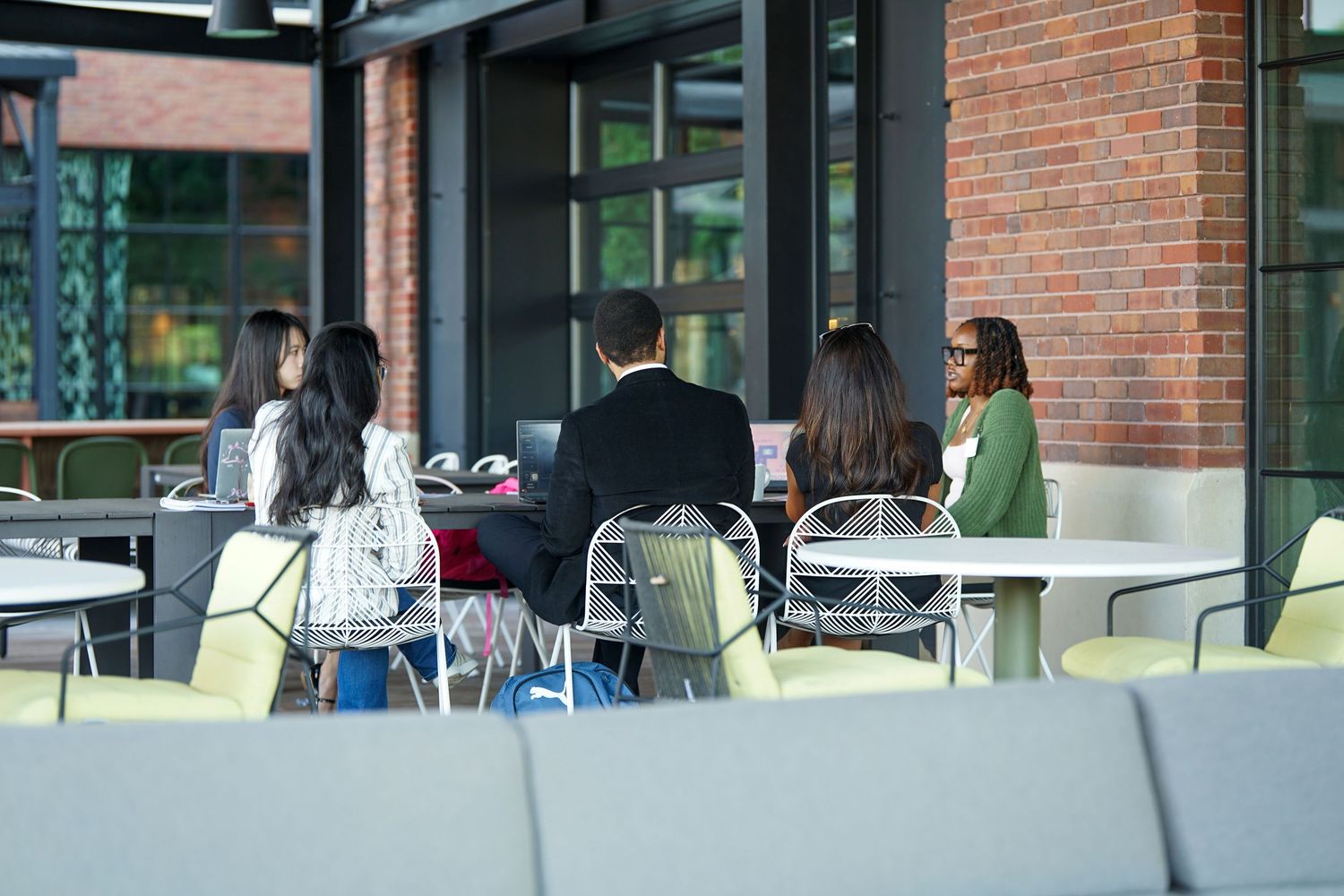 Students sitting at outdoor patio tables near a brick building, engaged in a group discussion on campus.