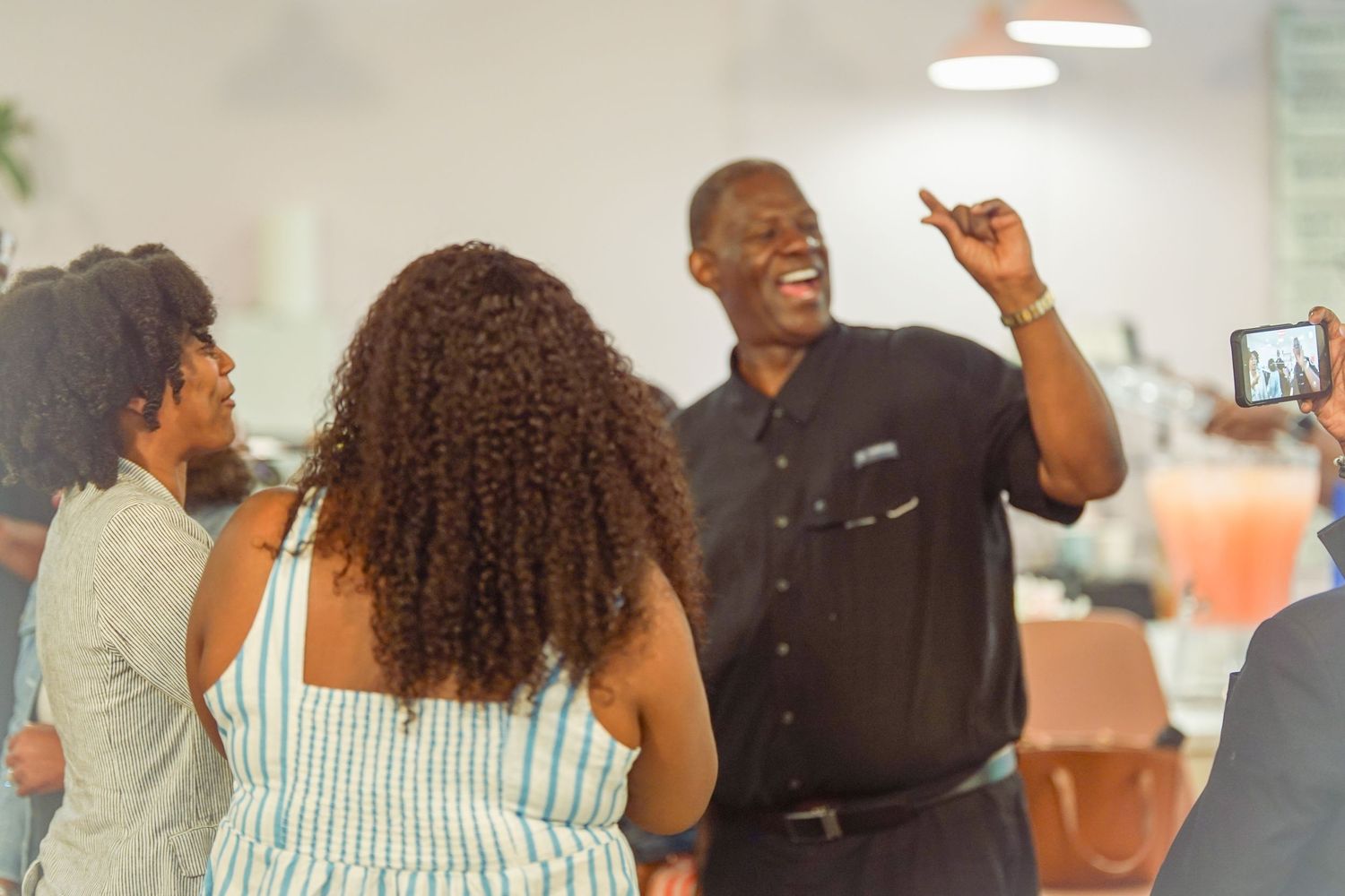 A smiling man in a black shirt gestures while speaking to a group of people at an indoor social gathering.