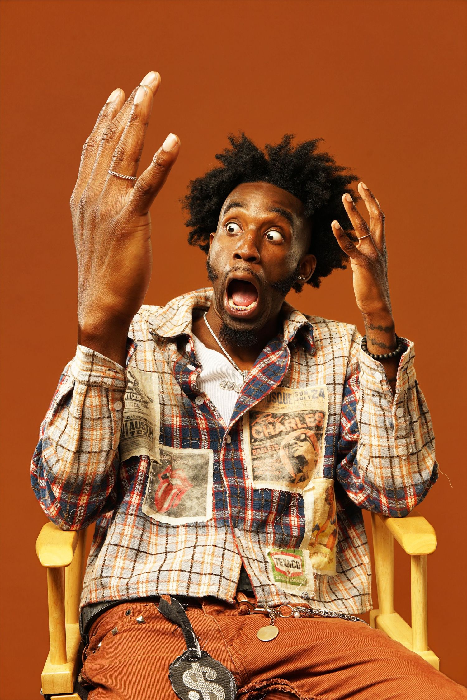 Expressive portrait with dramatic hand gestures in casual patchwork outfit against orange background.