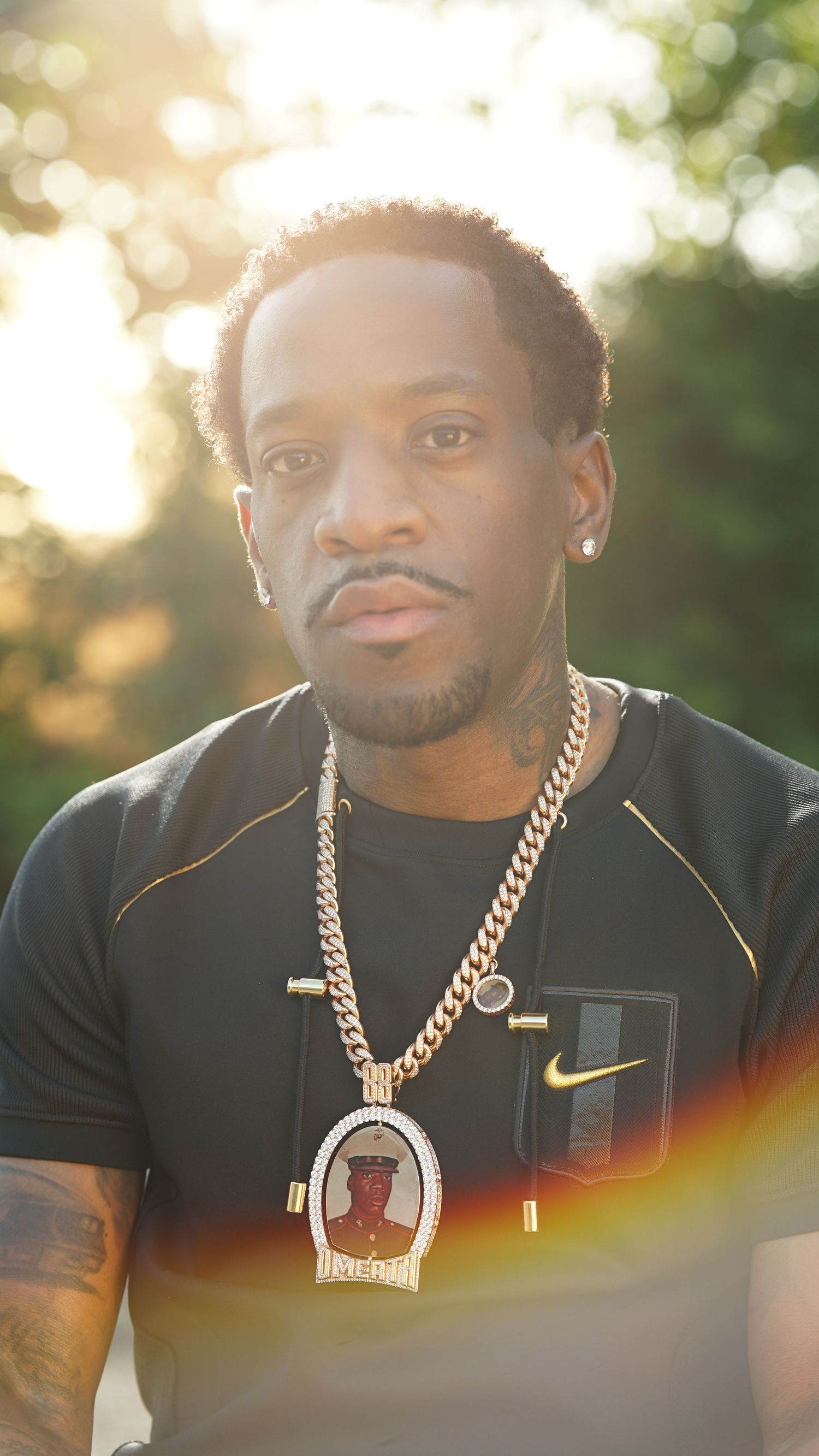Person wearing a black Nike shirt and gold chain necklace poses outdoors in natural lighting.