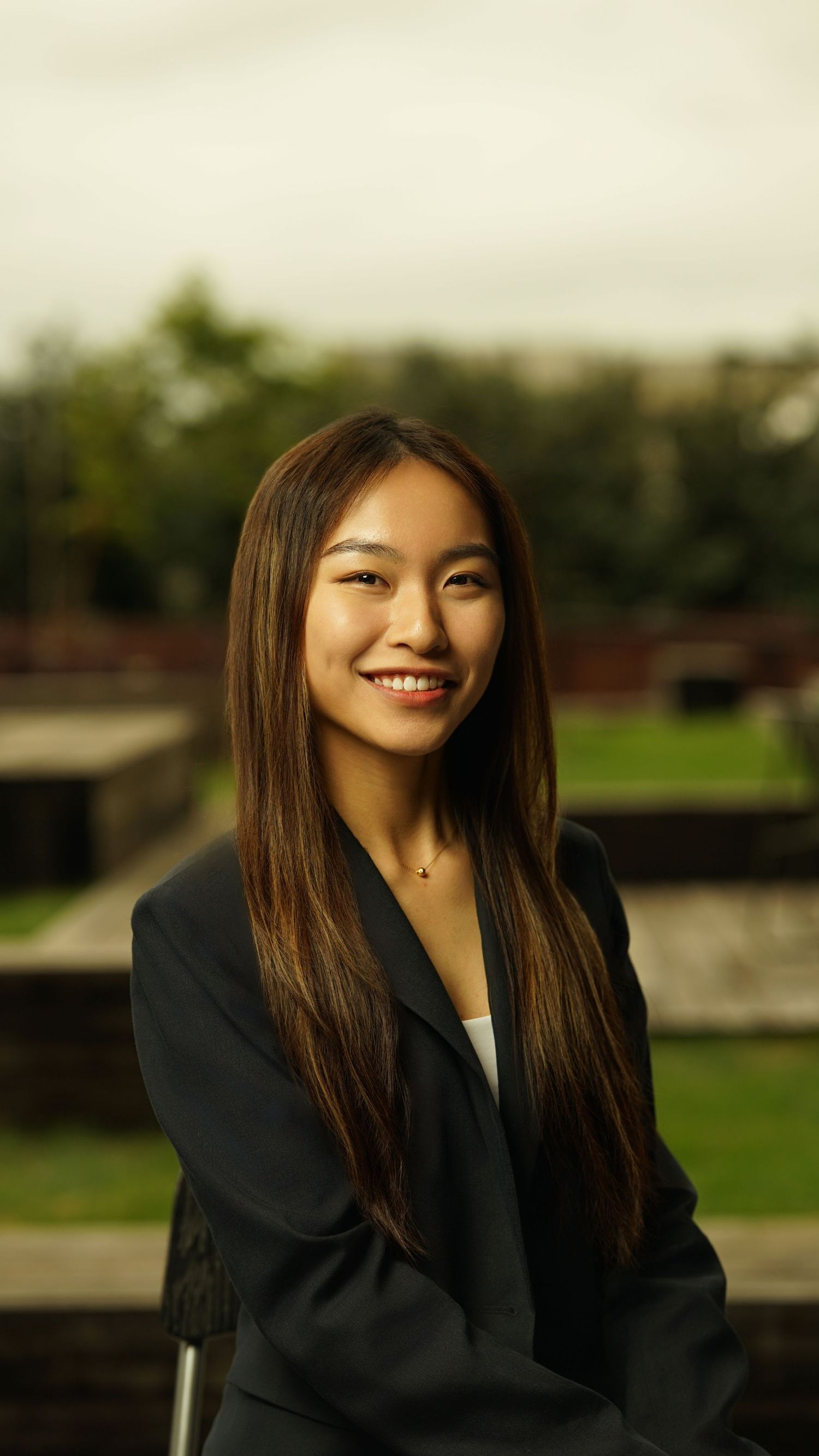 Professional portrait of person in dark blazer with long straight hair against outdoor setting.