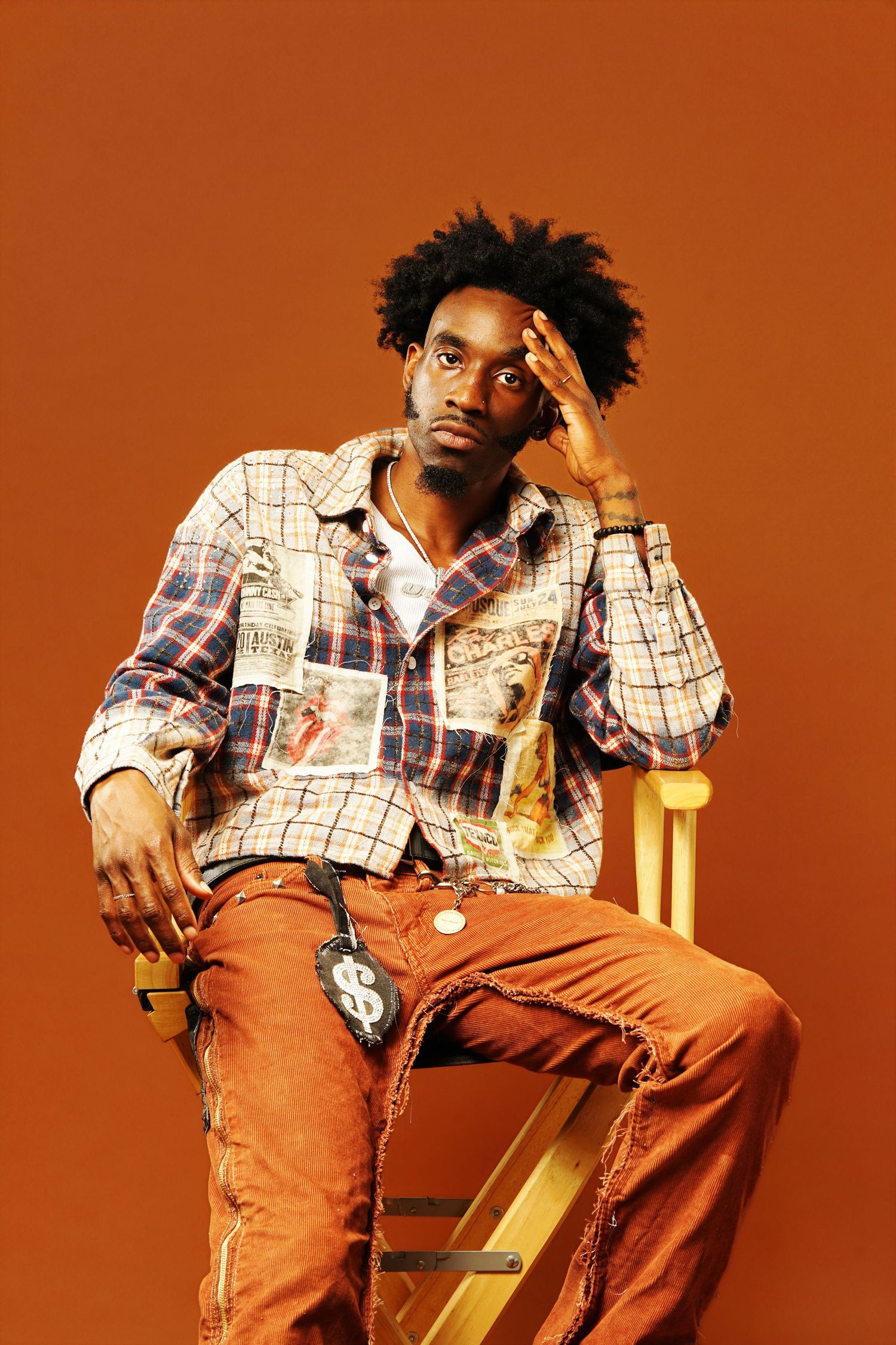 Stylish model in patchwork plaid jacket and orange pants poses thoughtfully against warm orange backdrop.