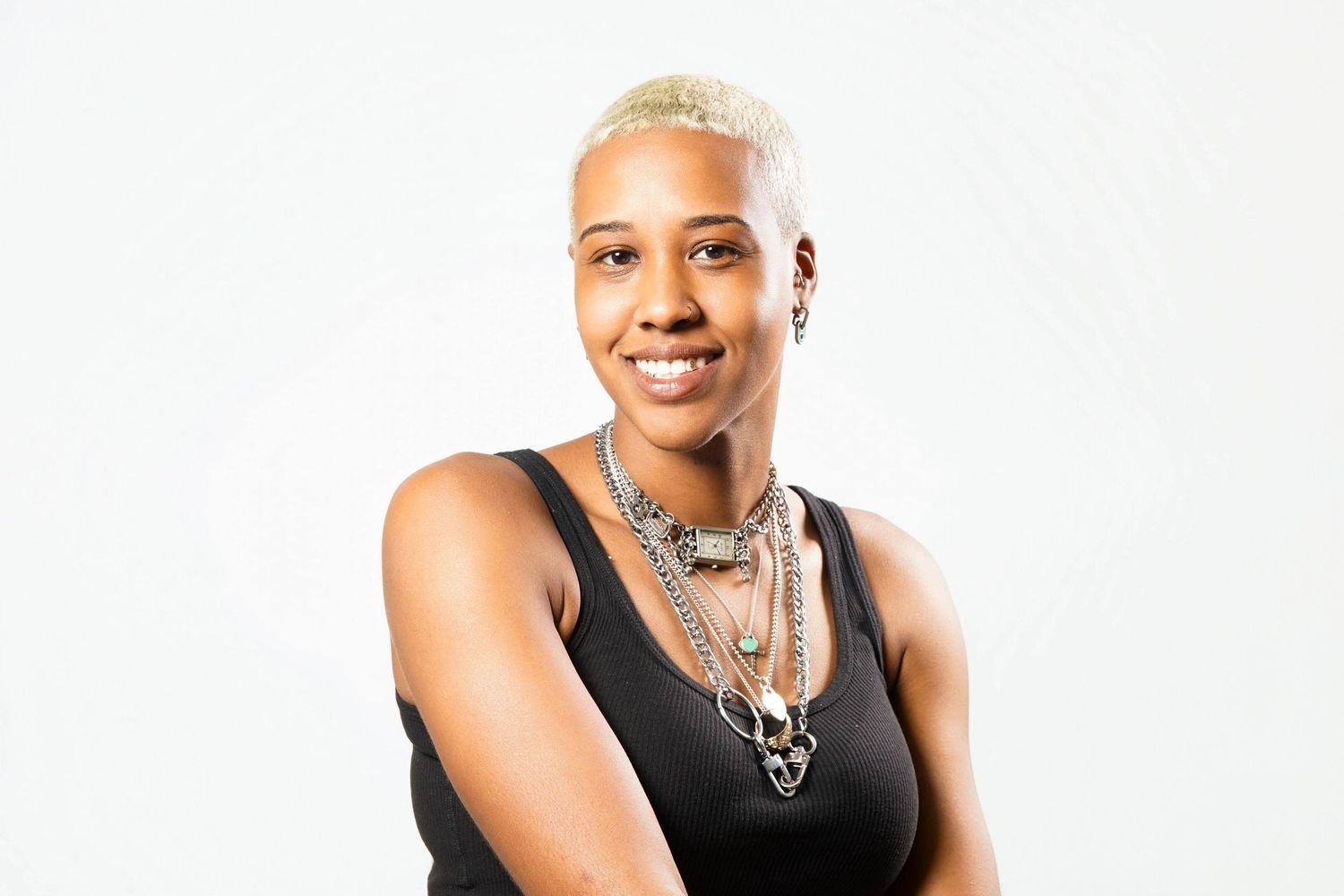 Smiling woman with blonde cropped hair wearing a black tank top and layered silver necklaces against a white background.