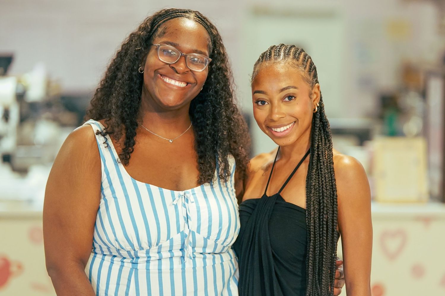 Two smiling women posing together indoors, one wearing glasses and a blue striped dress, the other in a black outfit.