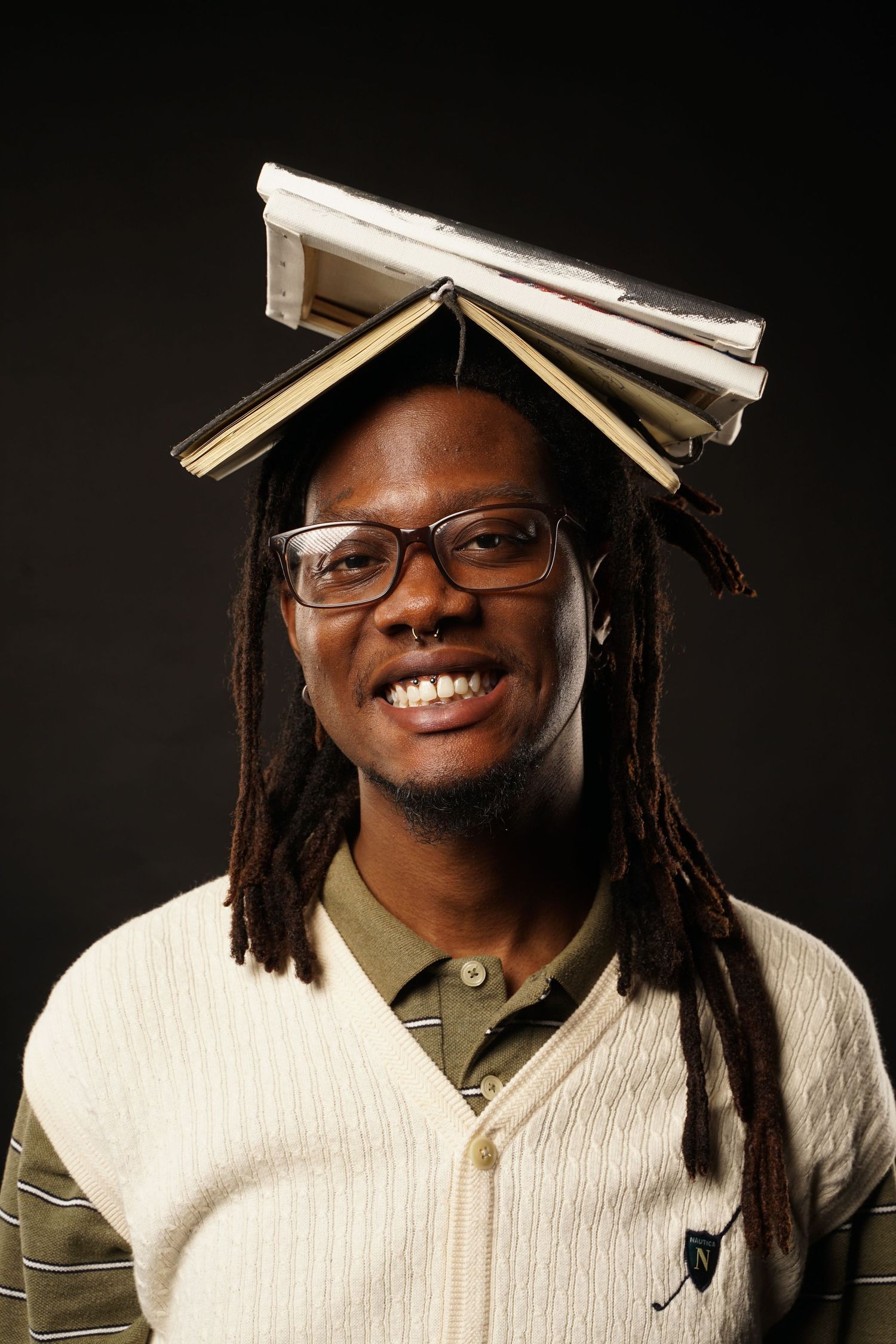 Person with dreadlocks and glasses balances books on their head while smiling against dark background.