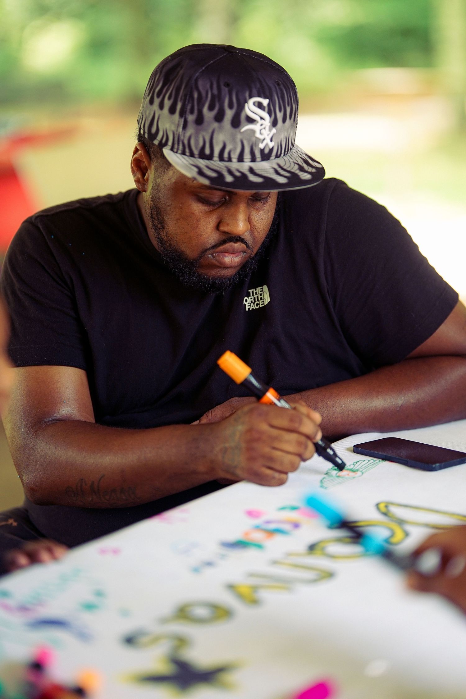 Man in Chicago White Sox cap writing colorful letters on a poster board with markers outdoors.