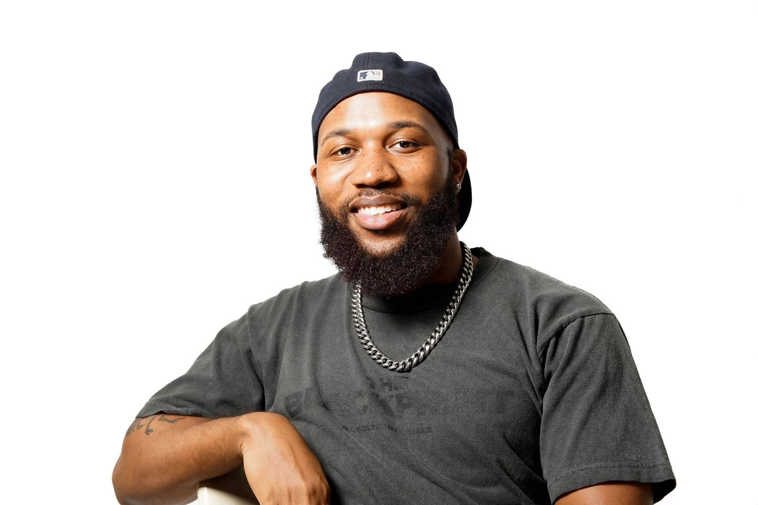 Smiling bearded Black man wearing a gray t-shirt, blue beanie, and silver chain necklace against a white background.