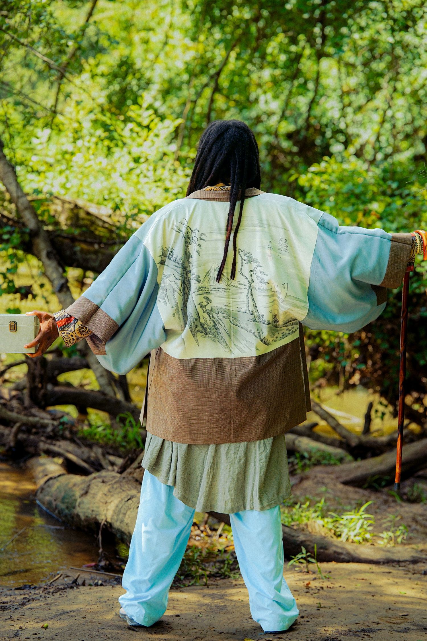 Person in decorative kimono jacket and light blue pants stands in a lush forest setting, viewed from behind.