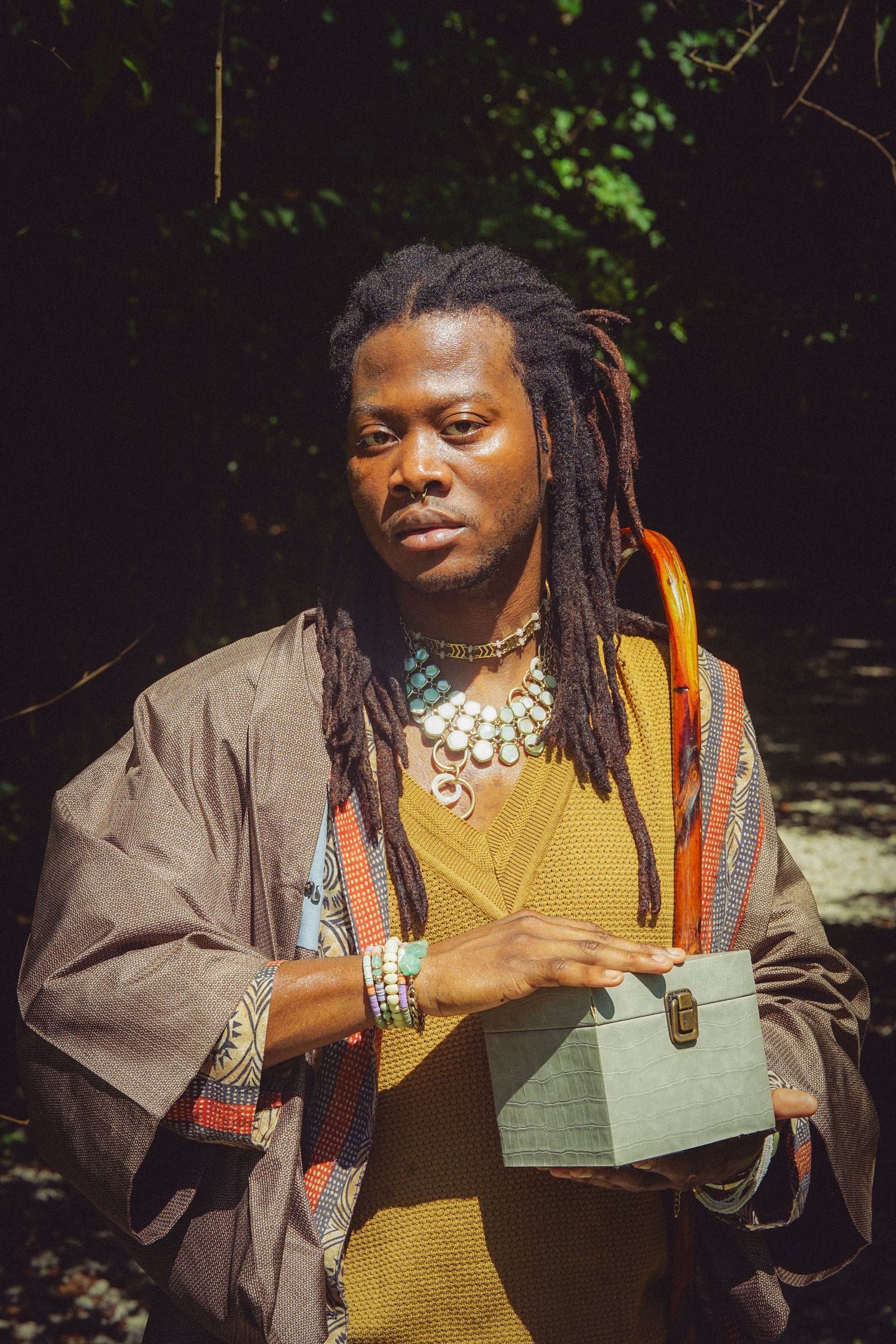 Man with dreadlocks wearing colorful jewelry and robes holds a small box outdoors in a forest setting.