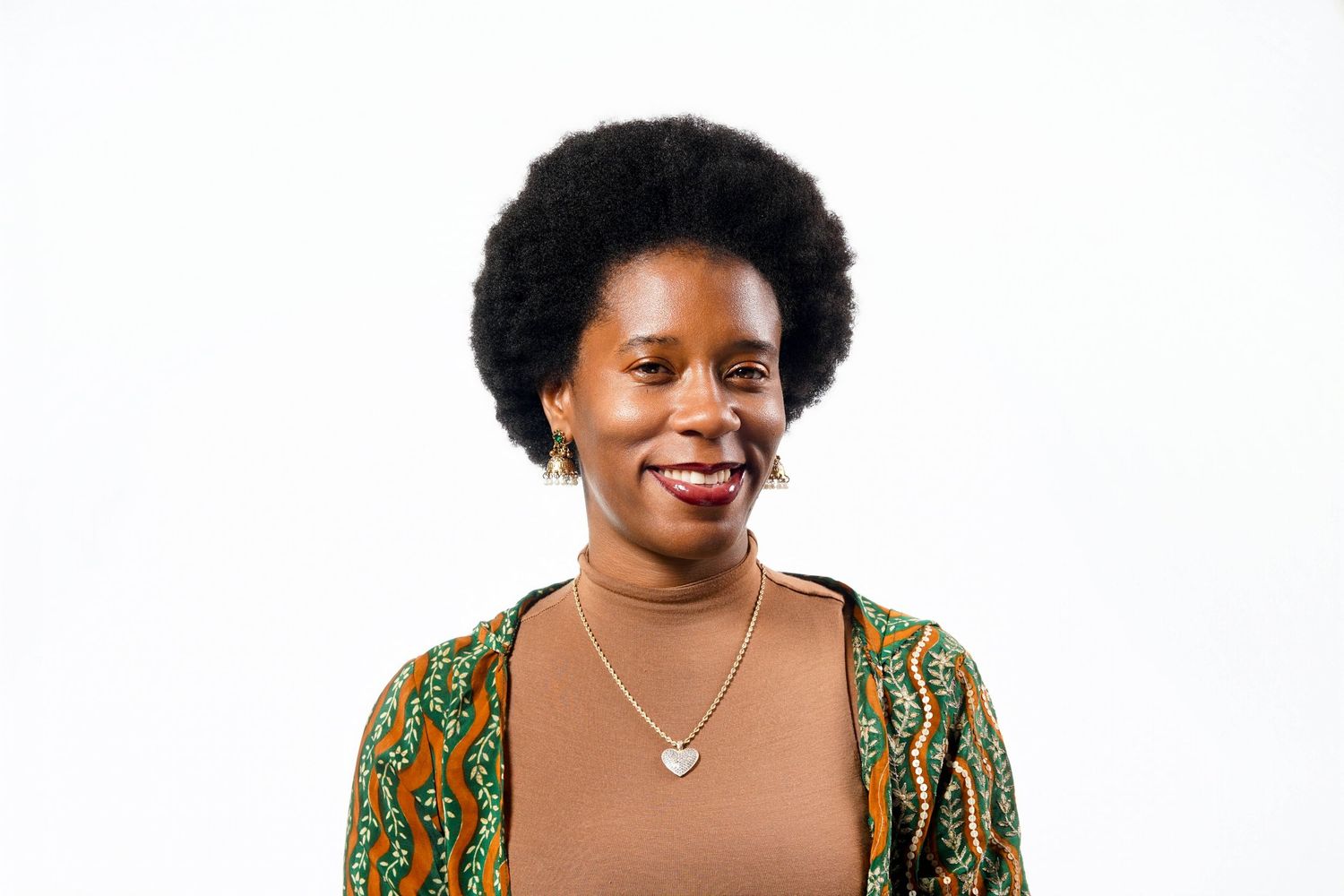 Smiling Black woman with natural afro hair wearing a green patterned cardigan and heart necklace against white background.