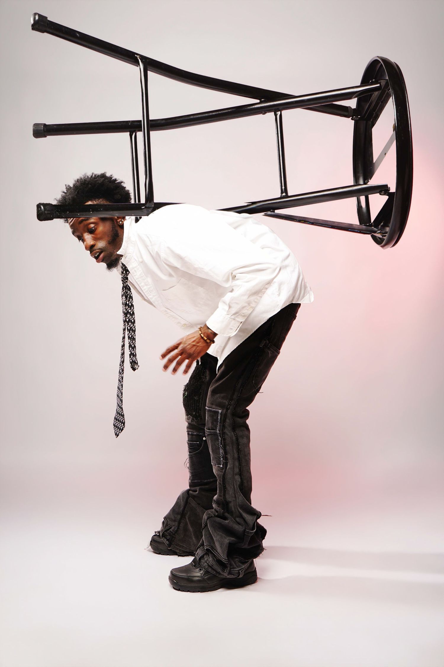 A man in a white shirt and tie balances a black chair on his head while bending forward in a studio.