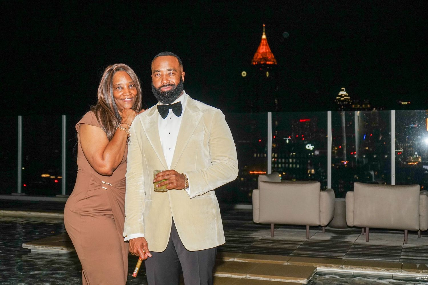Elegant couple posing on a rooftop at night with city skyline and illuminated tower in background.