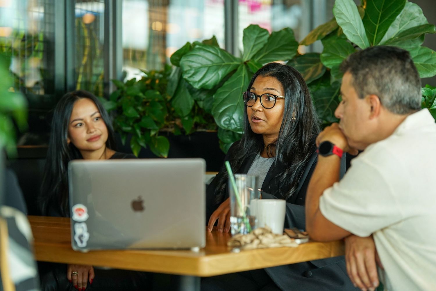 Three professionals collaborating at a cafe table with an Apple MacBook laptop surrounded by tropical plants.