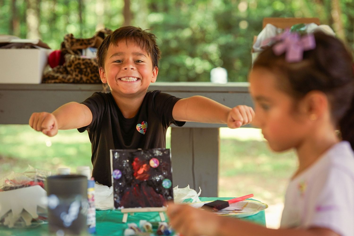 Smiling boy proudly displays colorful artwork at outdoor craft table with girl nearby.