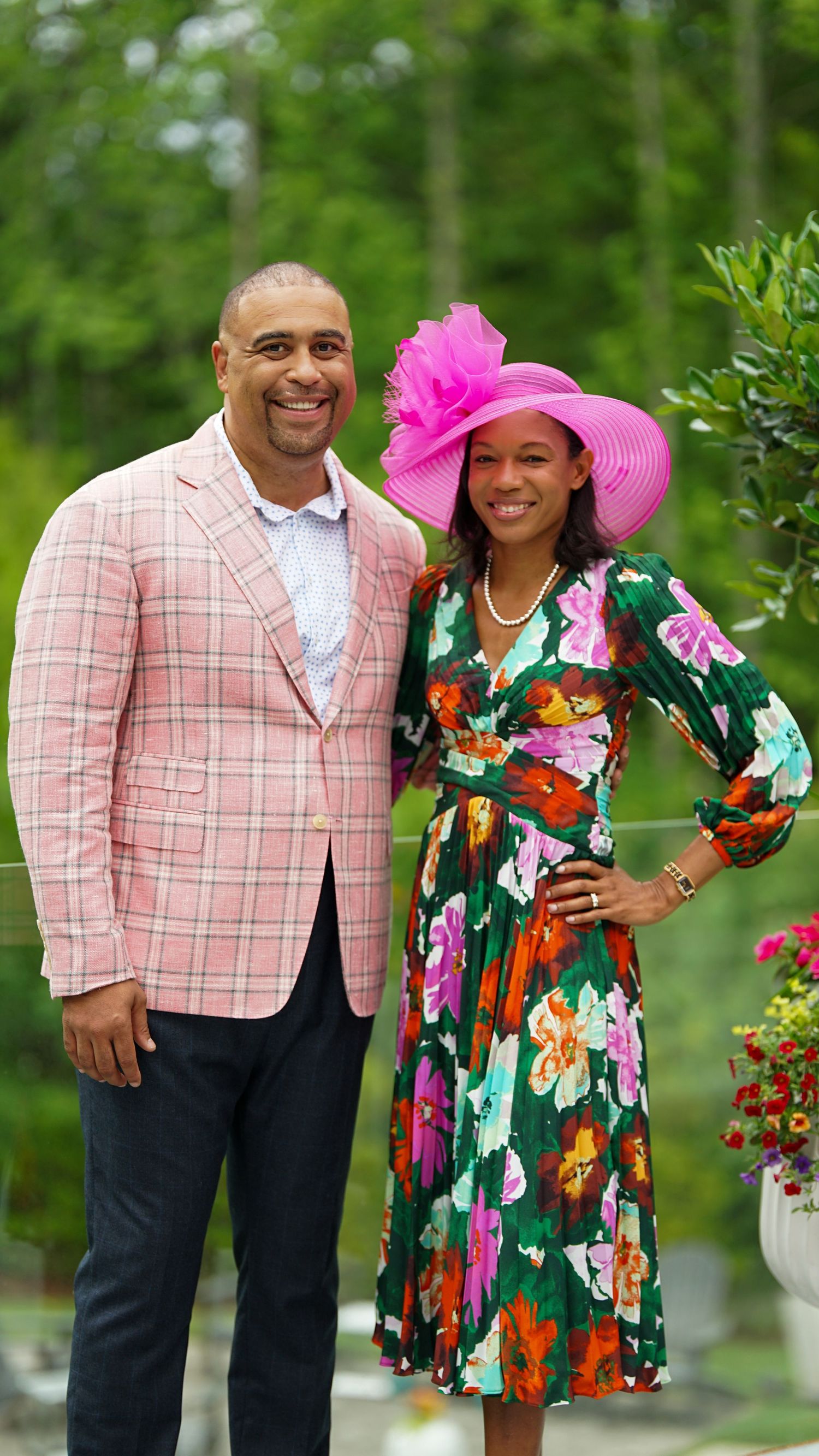 A couple poses outdoors in coordinated outfits with a pink plaid blazer and floral dress against a green garden backdrop.