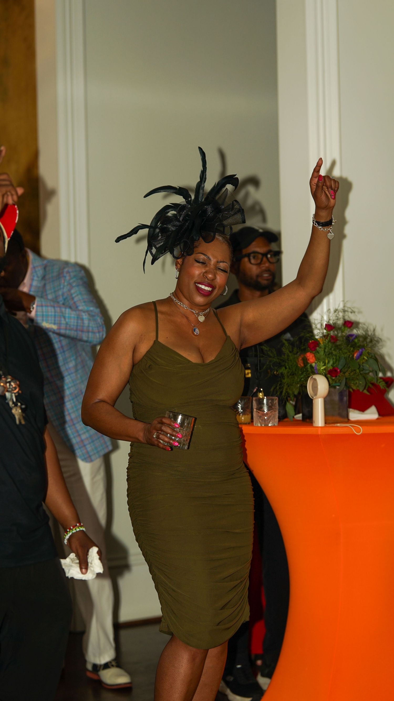 A person in an olive green dress dances energetically at an indoor party near an orange accent table.
