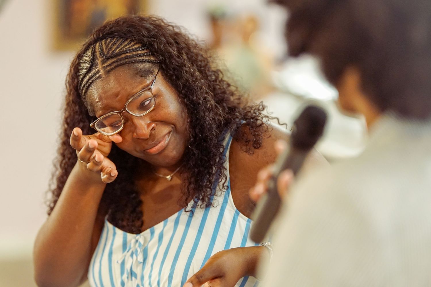 A woman wearing glasses wipes a tear while smiling emotionally during a heartfelt moment.