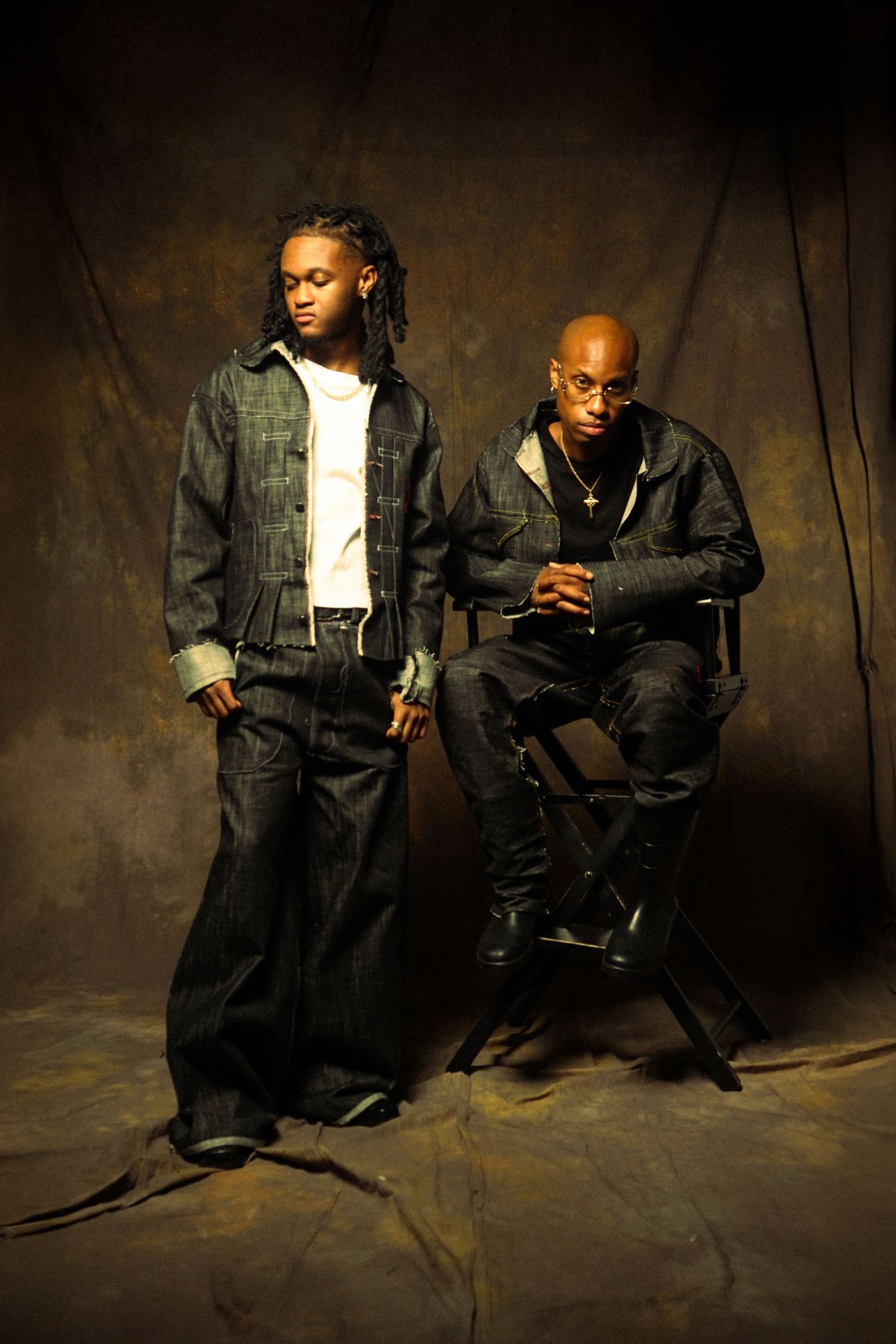 Dramatic studio portraits of two people in denim outfits sitting on chairs against a dark textured backdrop.