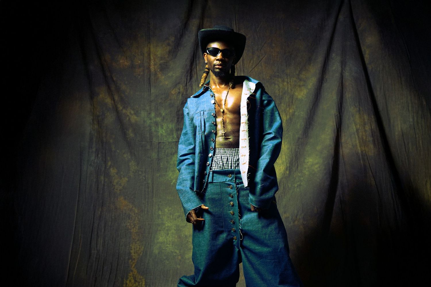 Person in blue denim jacket and sunglasses poses against dark textured background in moody lighting.