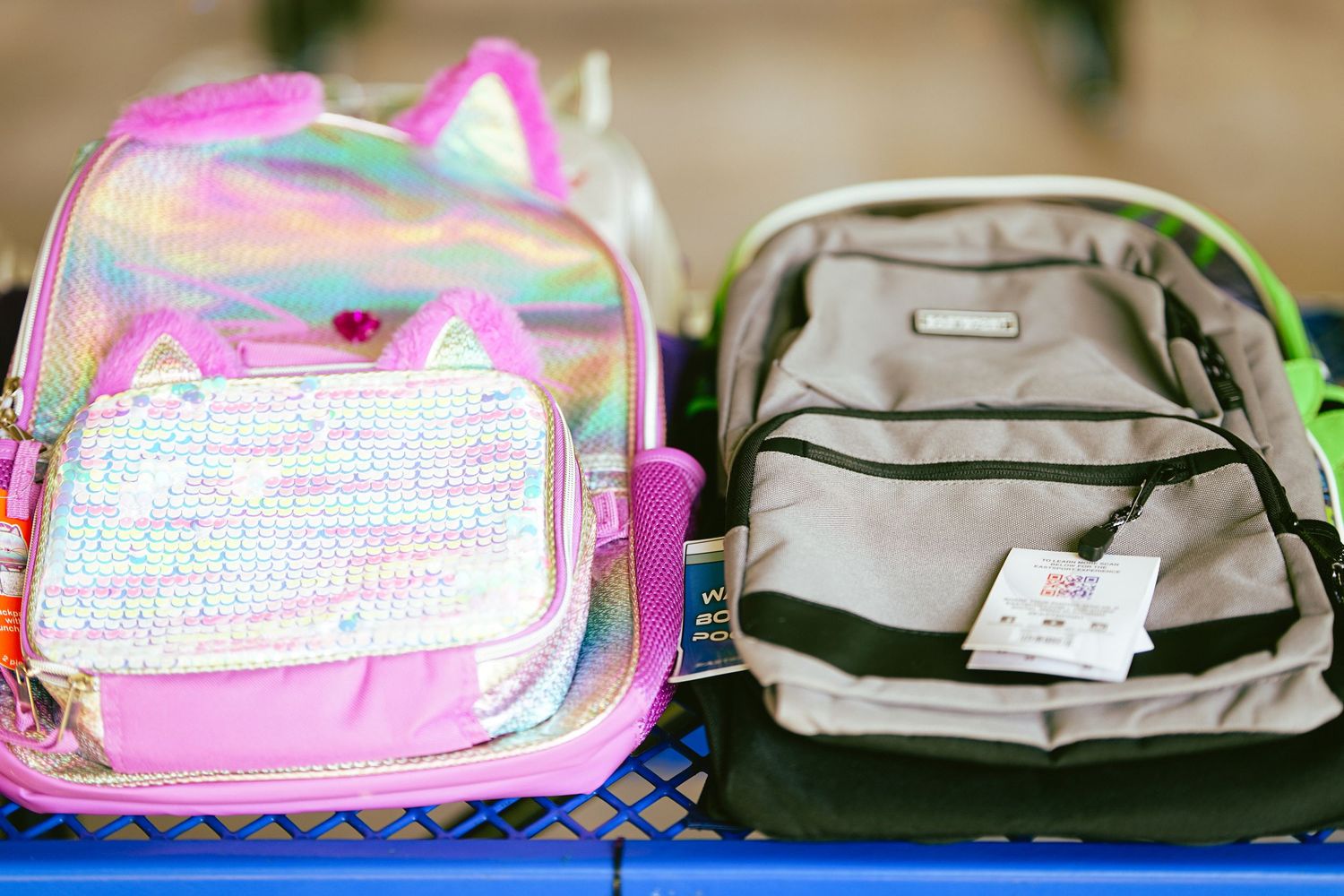 Two colorful backpacks on a blue cart, featuring a pink iridescent cat backpack and a green bag with price tag.