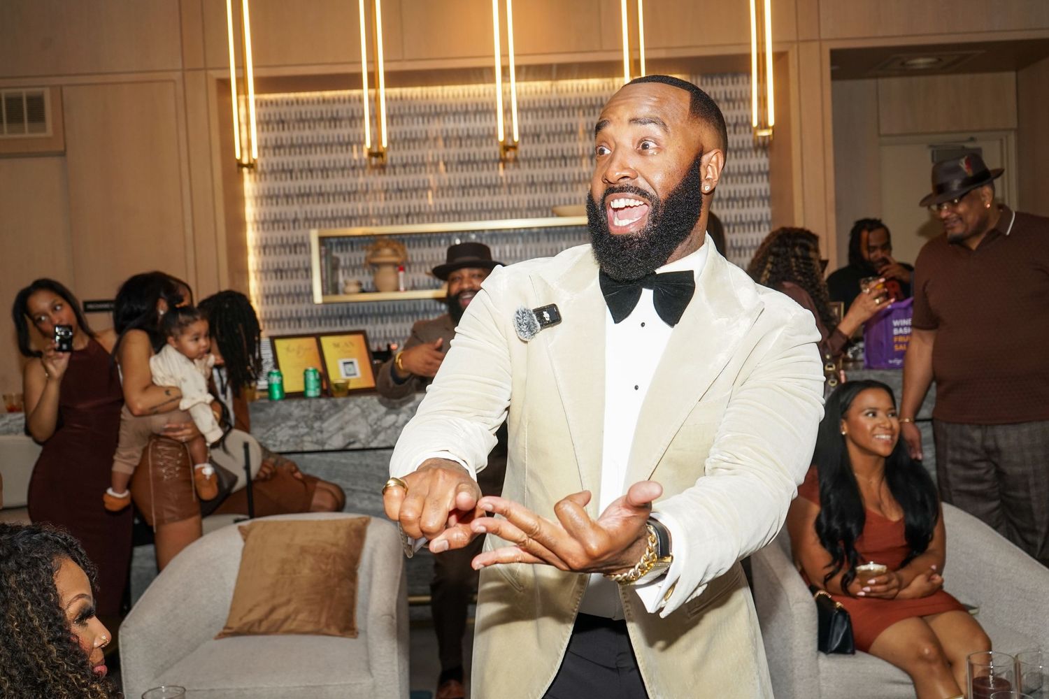 Man in cream tuxedo with black bow tie gestures at an elegant indoor party with guests in background.