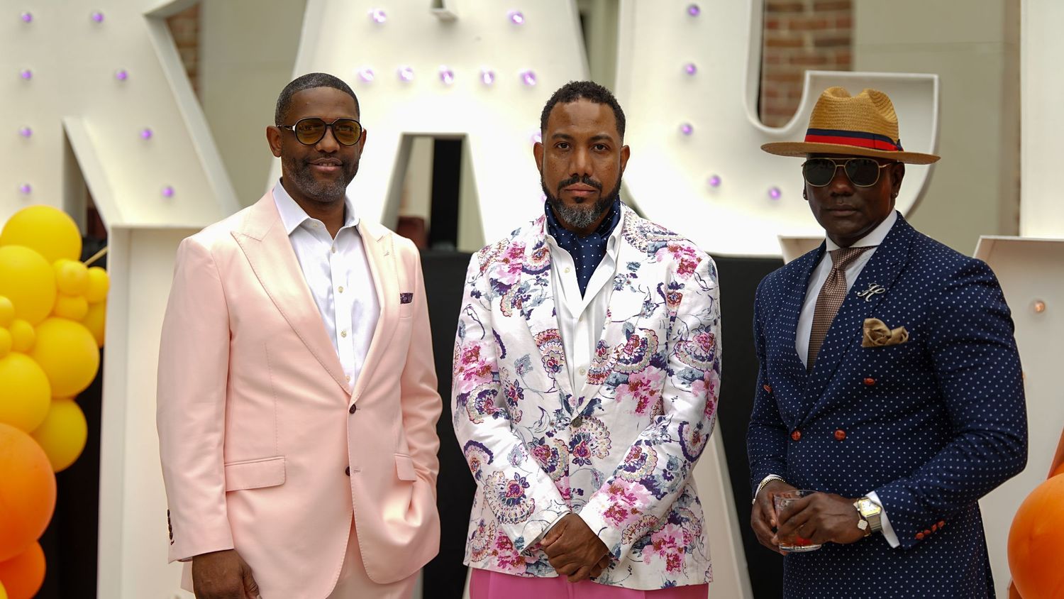 Three stylishly dressed men in formal suits pose together at an event with balloon decorations.