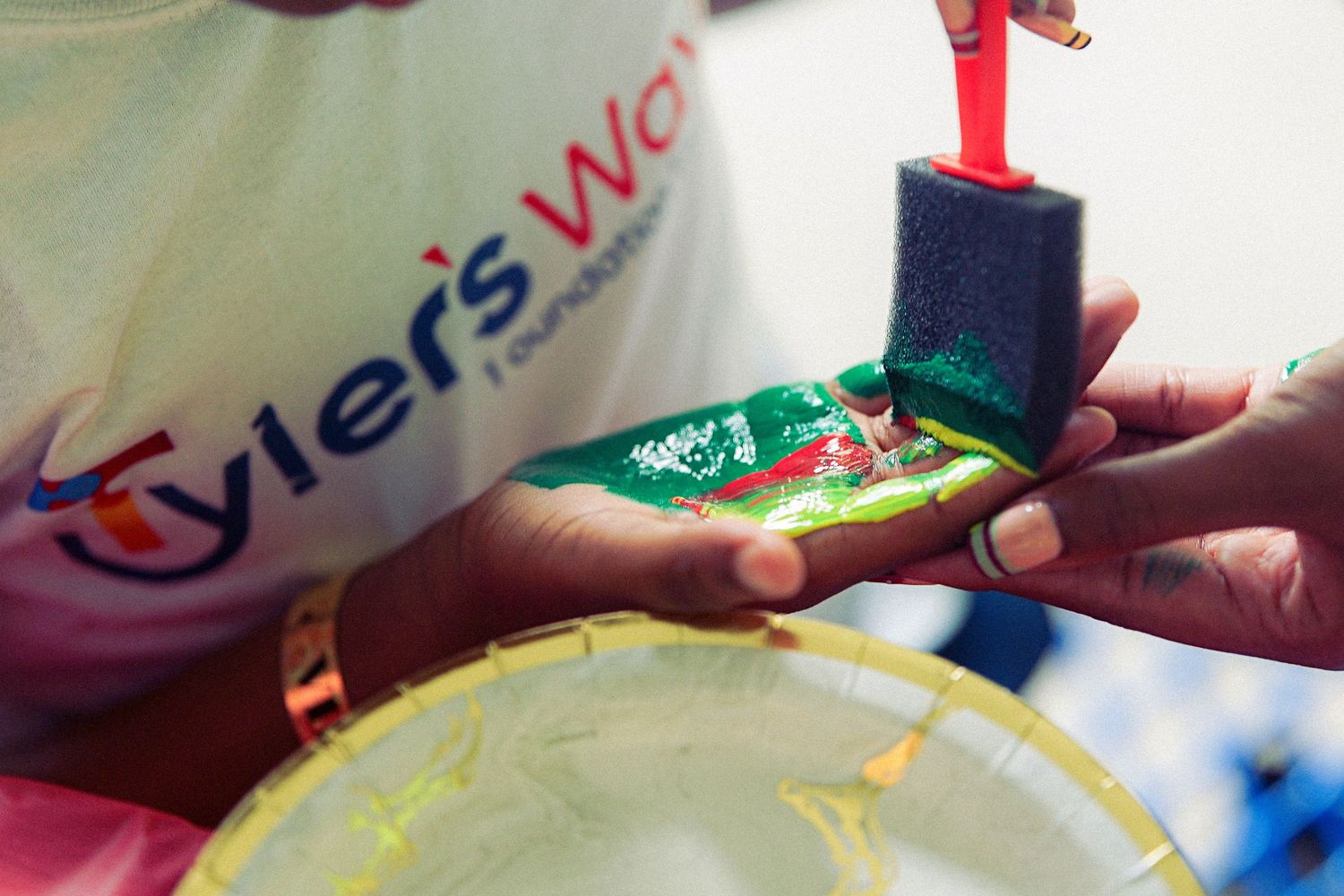 Hands holding a green foam sponge with red paint at a Tyler's Way Foundation crafting event outdoors.