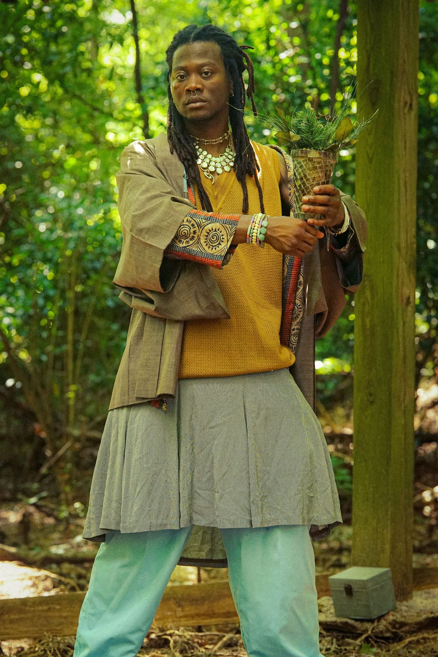 Man with dreadlocks in eclectic bohemian outfit holding a plant vessel in a lush green forest setting.