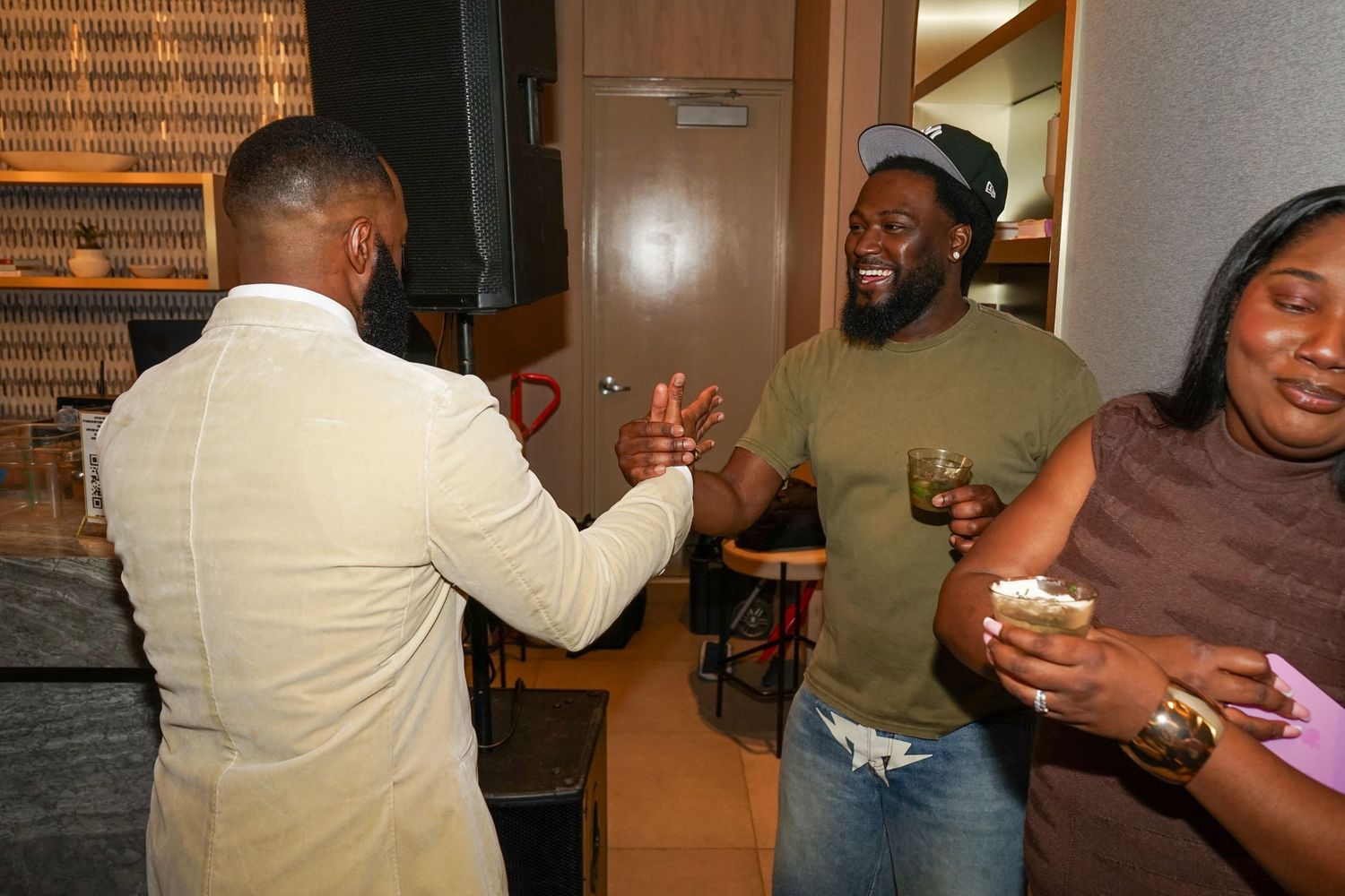 Three people socializing at an indoor gathering, laughing and holding drinks in a warmly lit venue.
