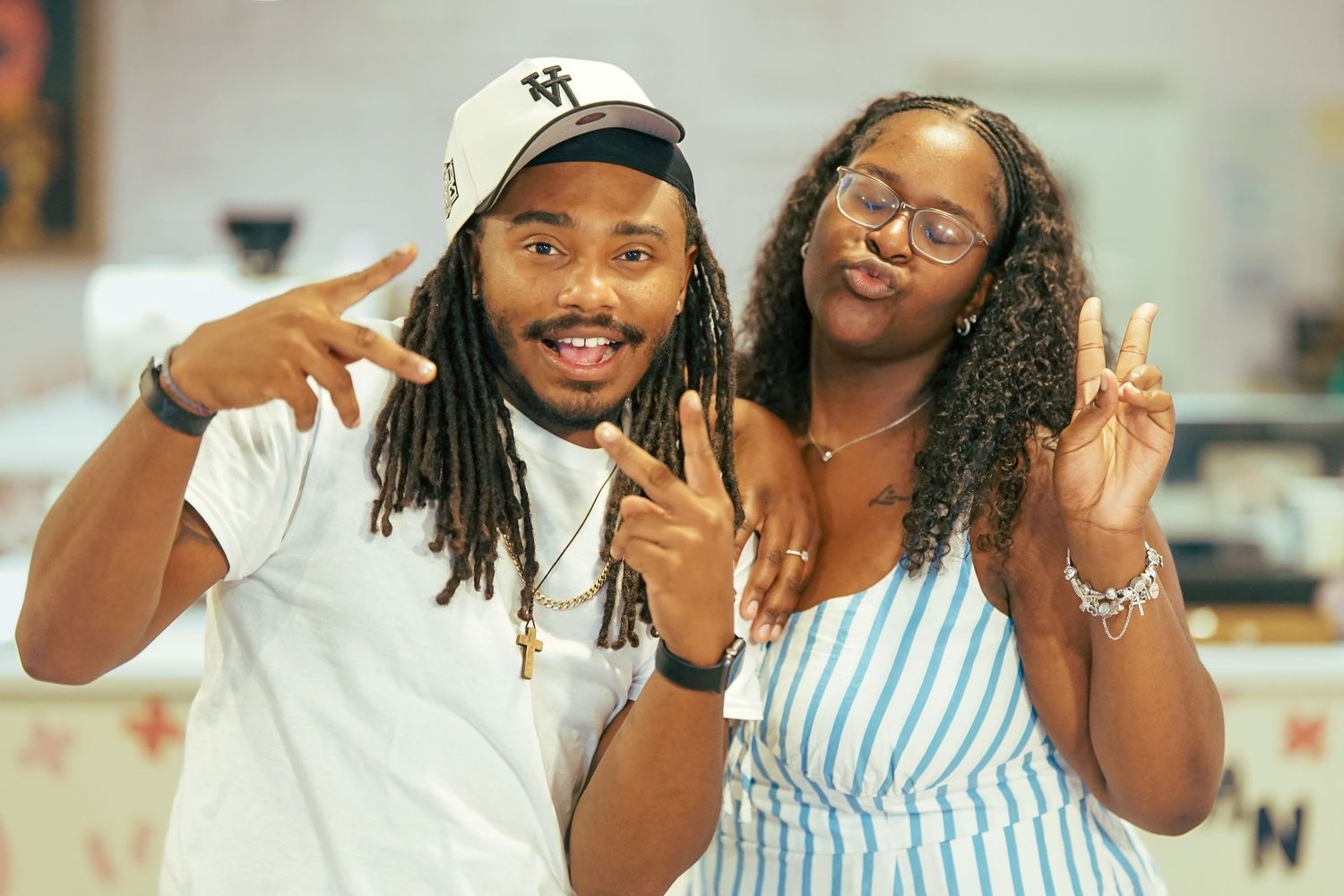 A young Black man and woman smile and make peace signs together at an indoor event.