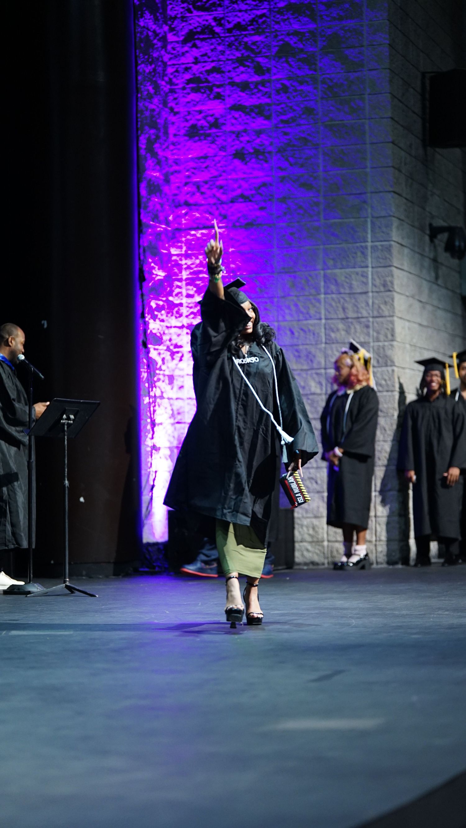 Graduates in black caps and gowns walk across a stage with dramatic purple lighting during commencement ceremony.