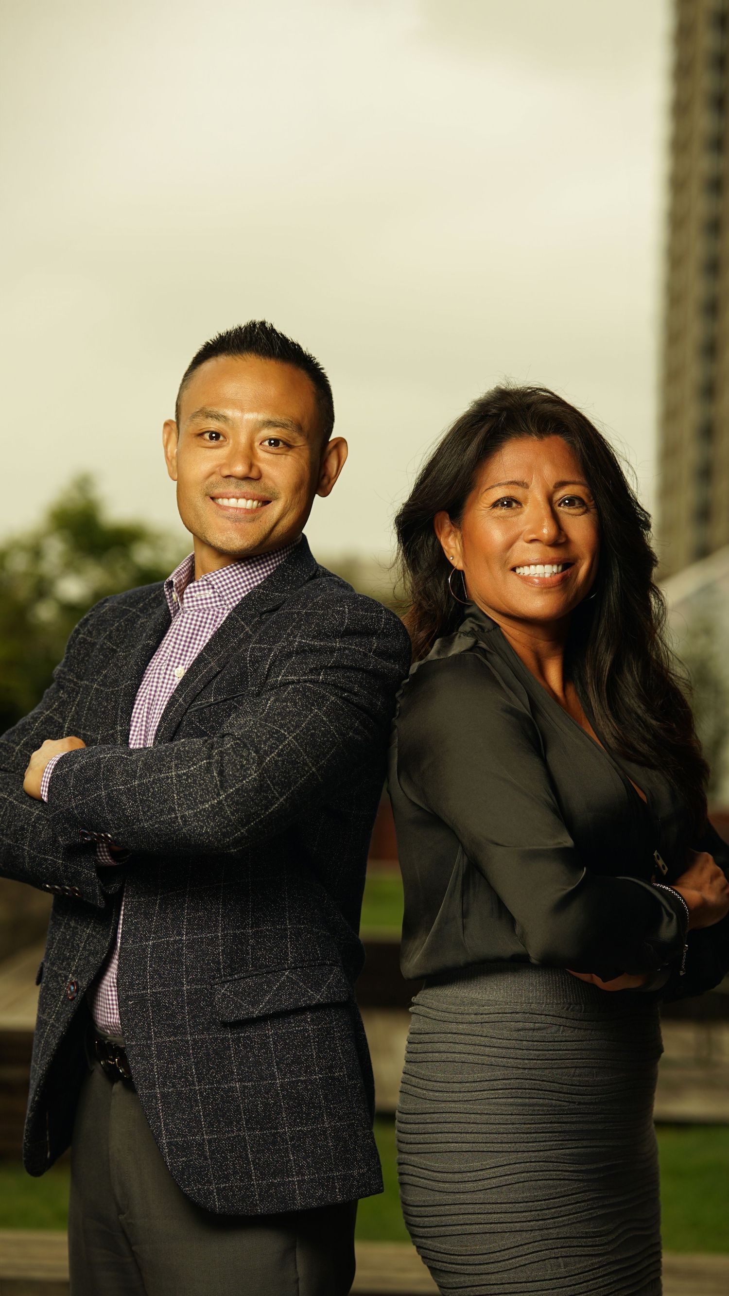 Two people posing together in professional attire with warm smiles against a blurred outdoor setting.