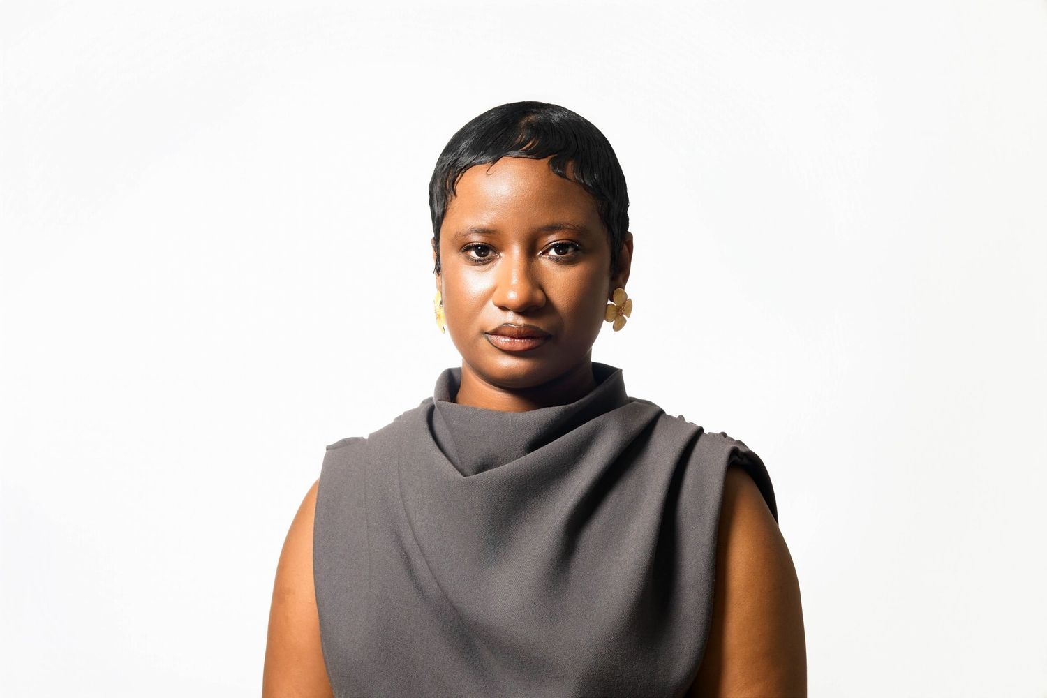 Young Black woman in gray sleeveless turtleneck top with gold earrings poses against white background.